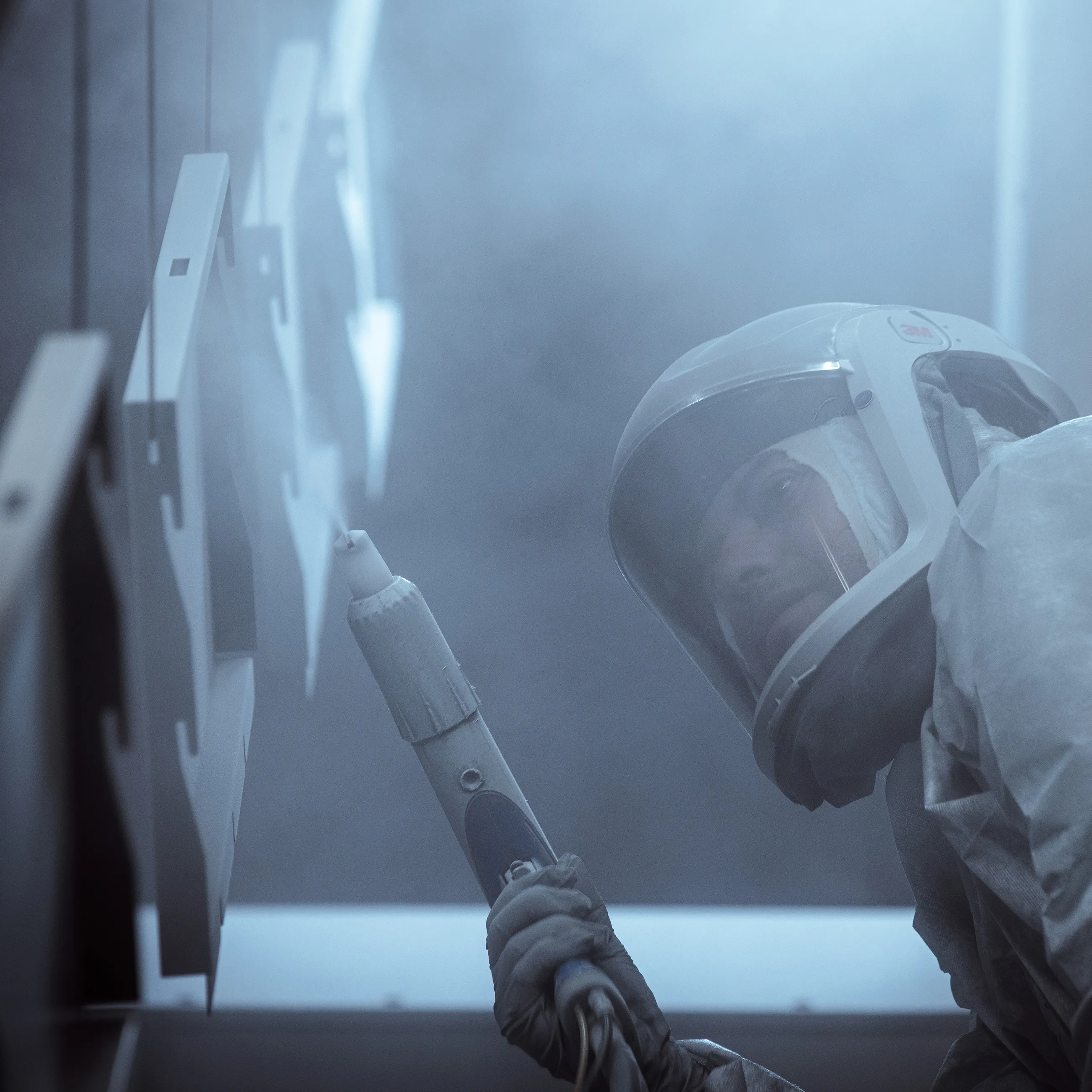 In an industrial setting with orange lighting, a person in protective gear showcases exceptional craftsmanship as they spray-paint metal frames, perfecting the art of custom modular cabinetry.