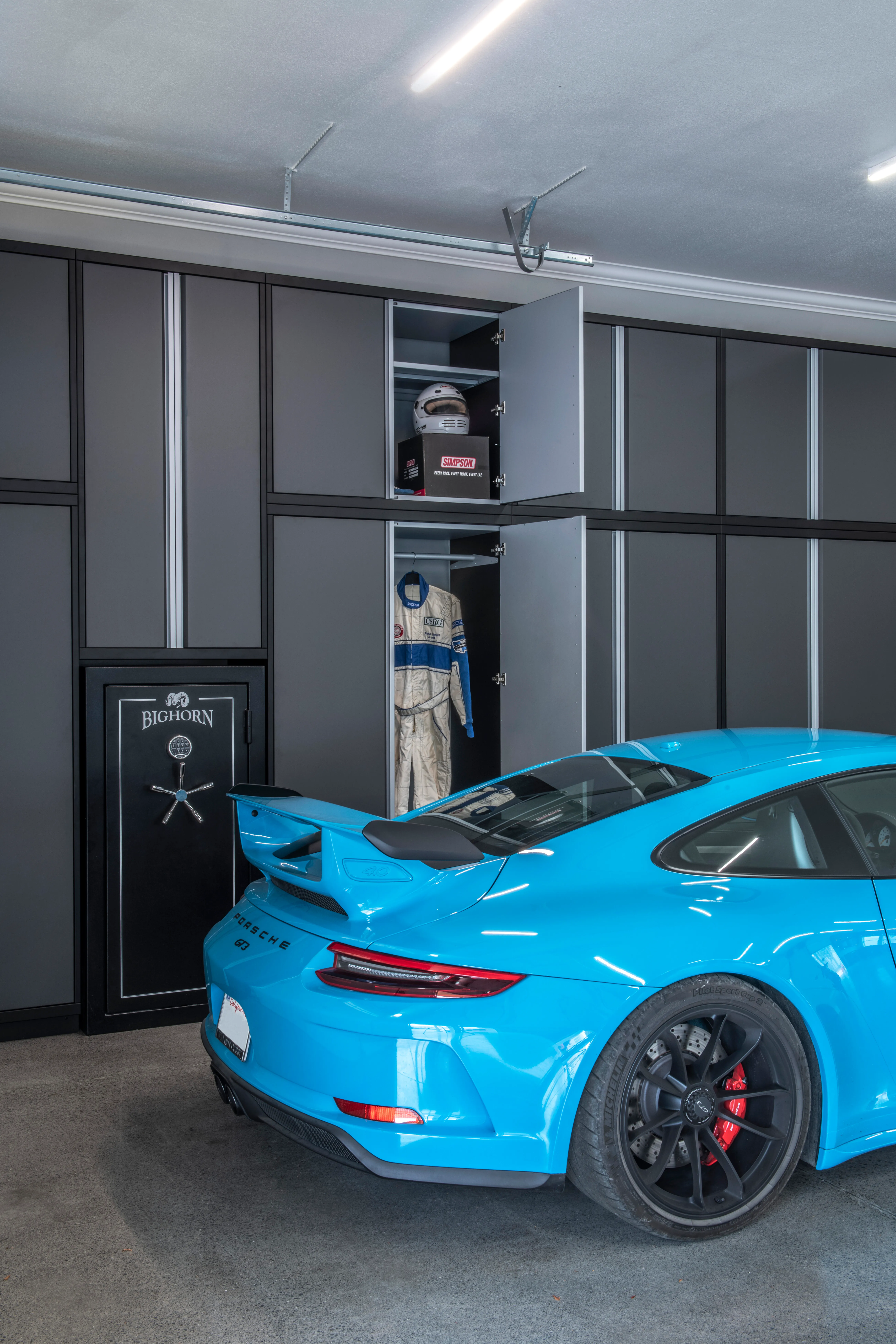 Luxury garage setup featuring custom matte gray Baldhead Cabinets with vertical storage for racing gear, Bighorn gun safe, and a vibrant blue Porsche 911 GT3, showcasing premium garage organization and performance lifestyle integration.