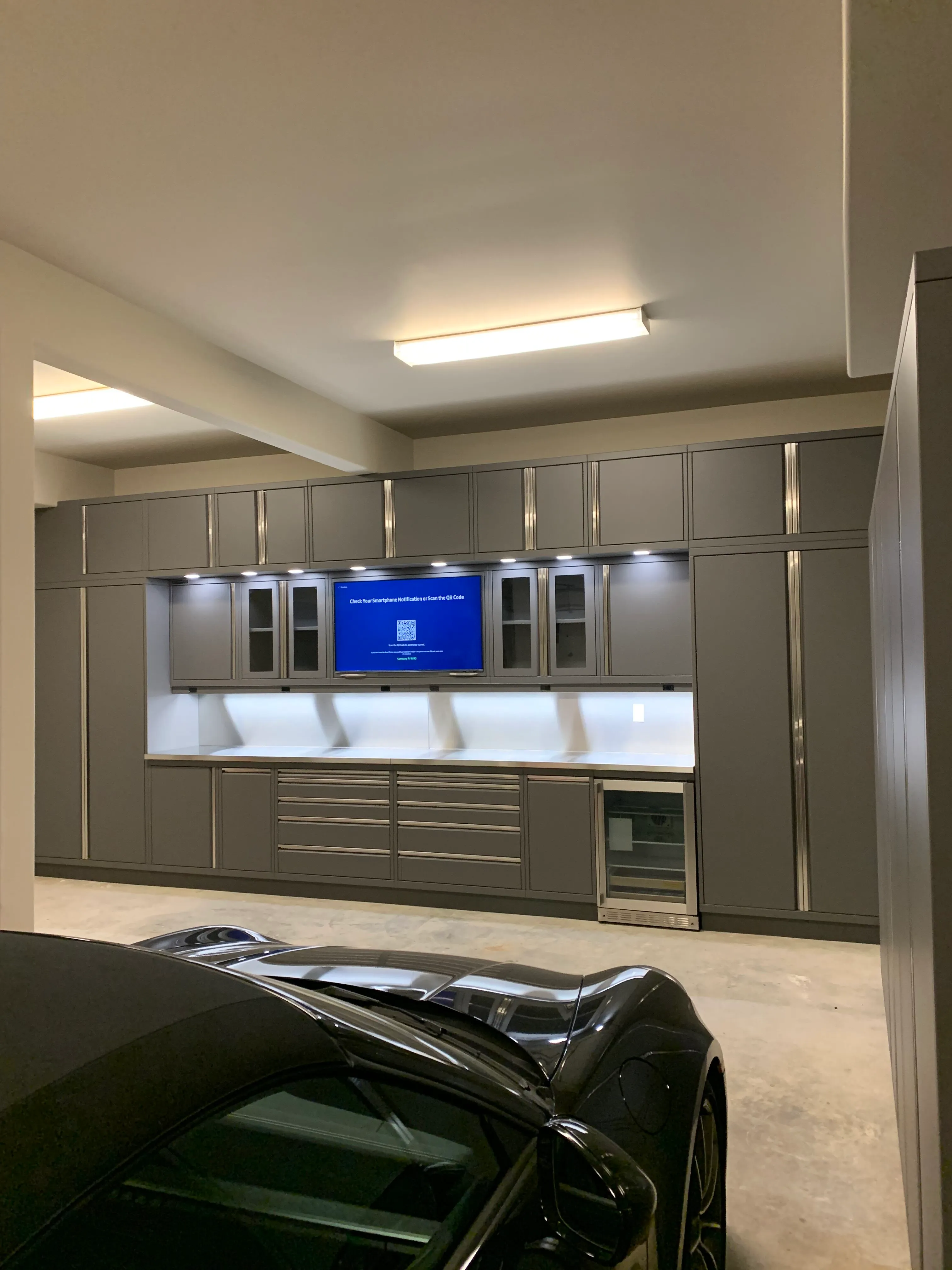 Custom luxury garage cabinetry by Baldhead Cabinets in matte gray with integrated LED lighting, glass display doors, under-counter beverage cooler, and flat-screen TV — designed for high-end automotive environments.