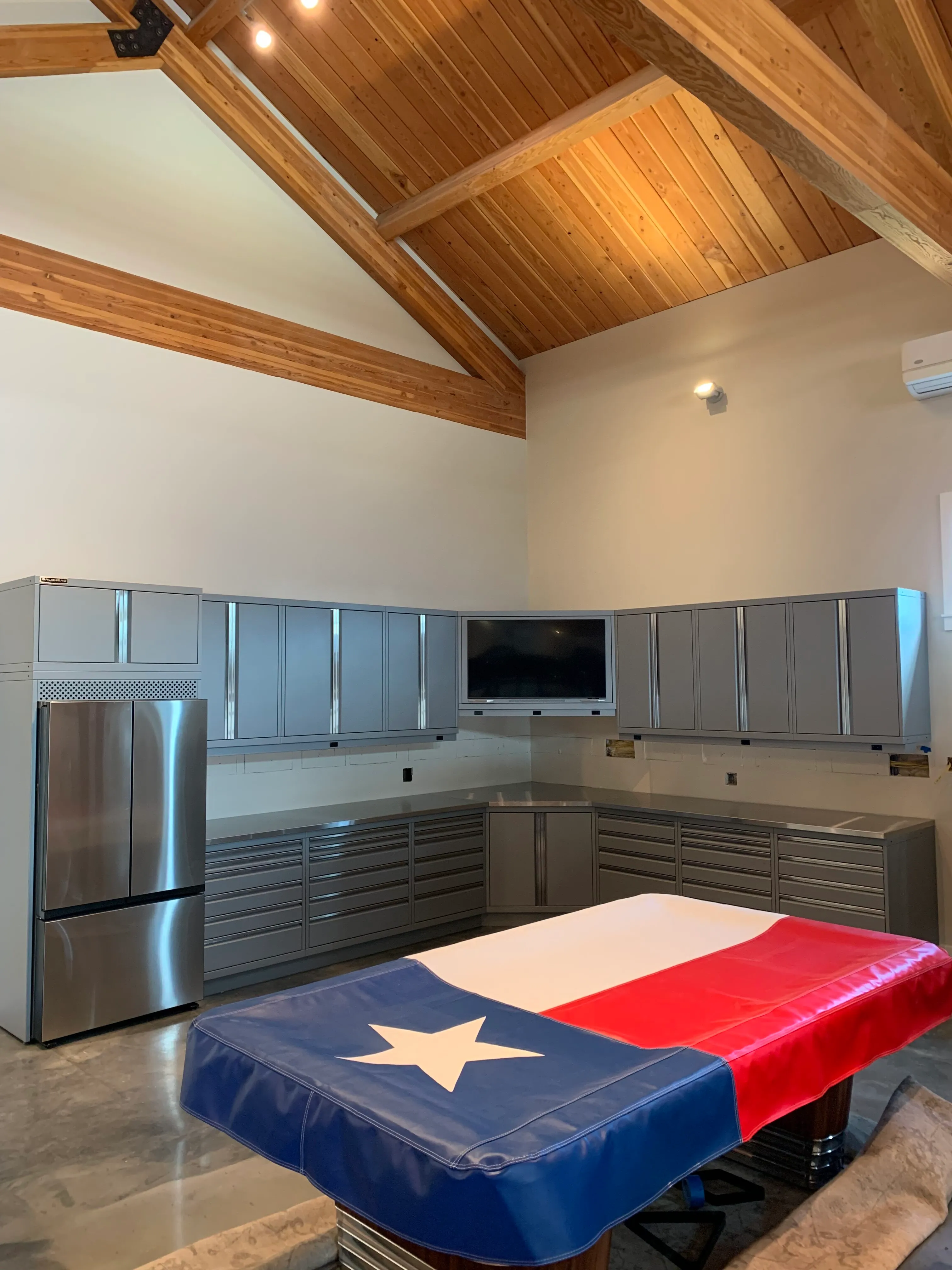 Modern gray garage cabinets with stainless steel countertops, integrated TV, and sleek drawer storage by Baldhead Cabinets — custom-built metal cabinetry for residential garages with vaulted wood ceilings and premium finishes