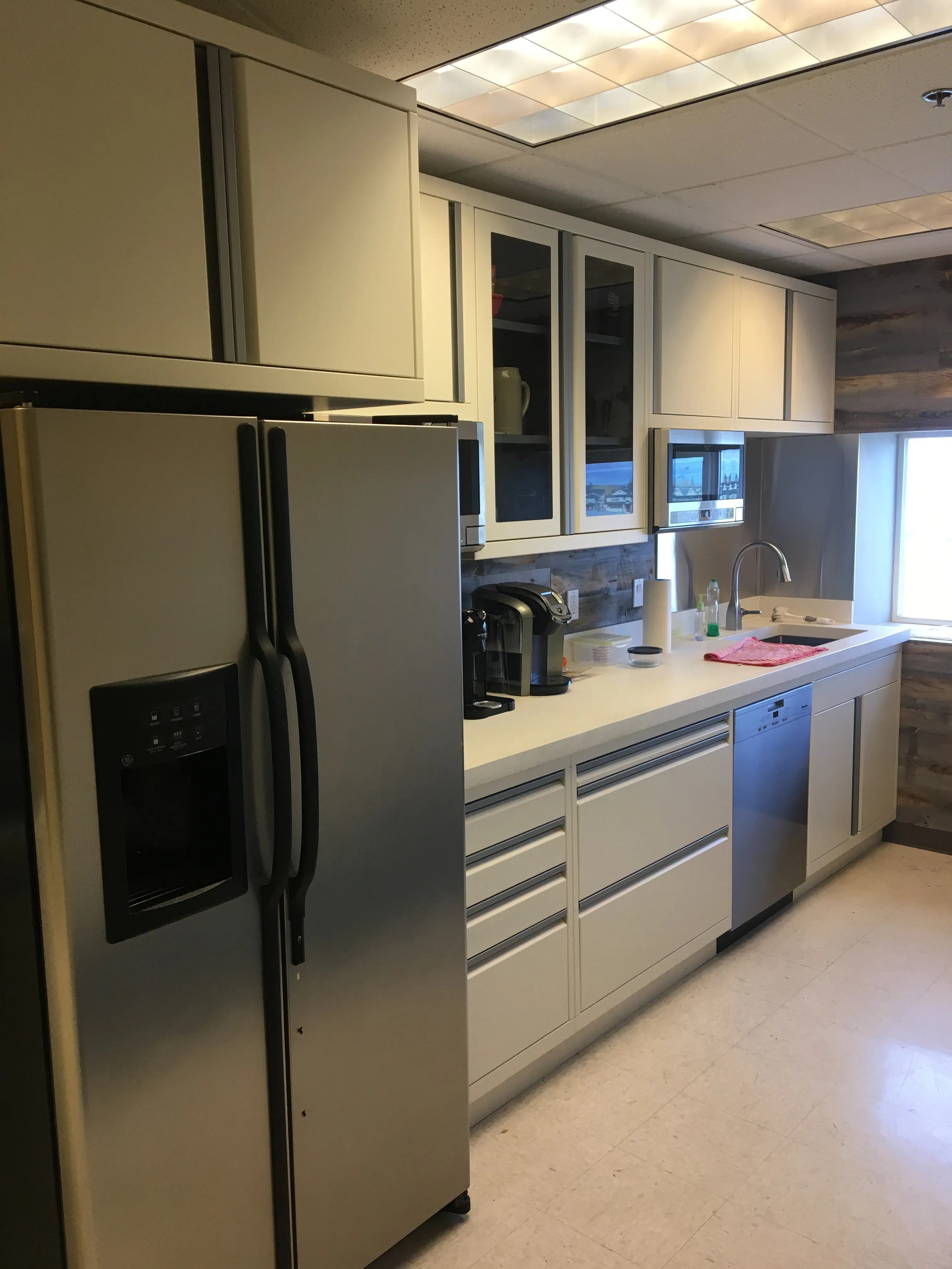 Modern kitchen installation by Baldhead Cabinets featuring white powder-coated cabinetry with integrated drawer pulls, glass-front upper cabinets, stainless steel refrigerator and dishwasher, solid surface countertops, and under-cabinet lighting in a professional breakroom setting.