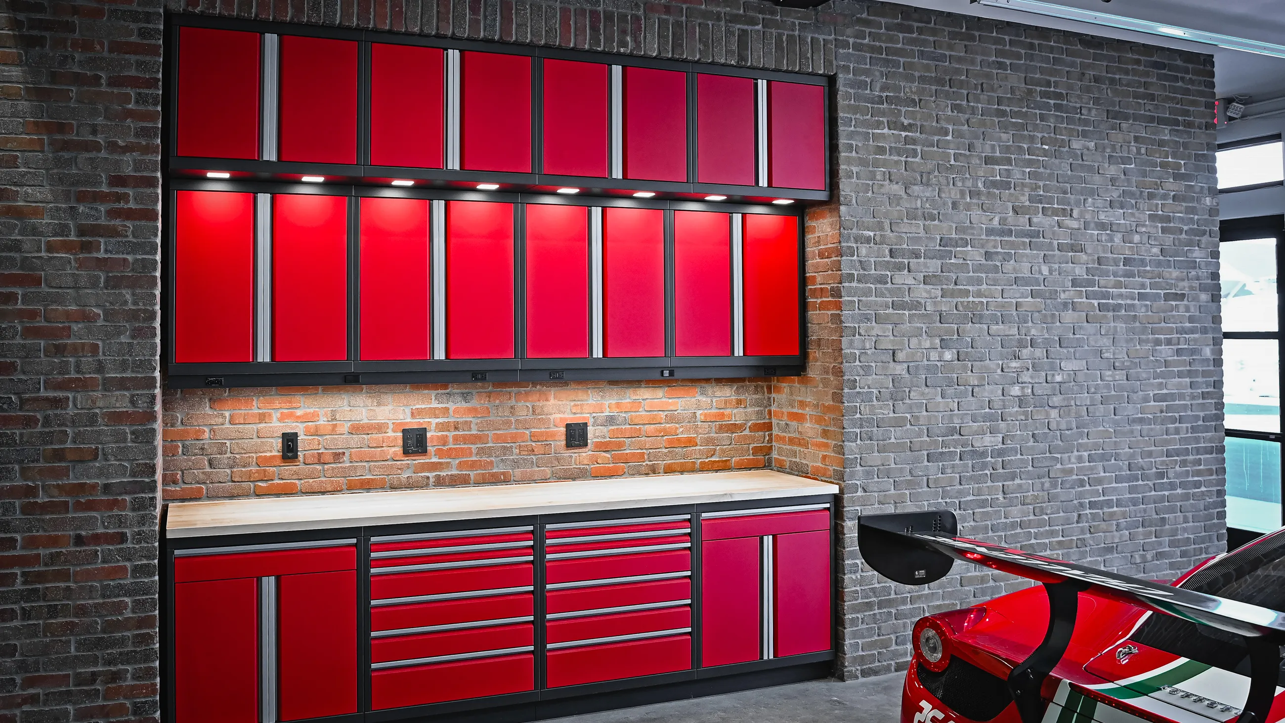 Bold red metal garage cabinets with LED task lighting and butcher block countertop by Baldhead Cabinets — custom storage solution set against an exposed brick wall in a high-performance automotive garage
