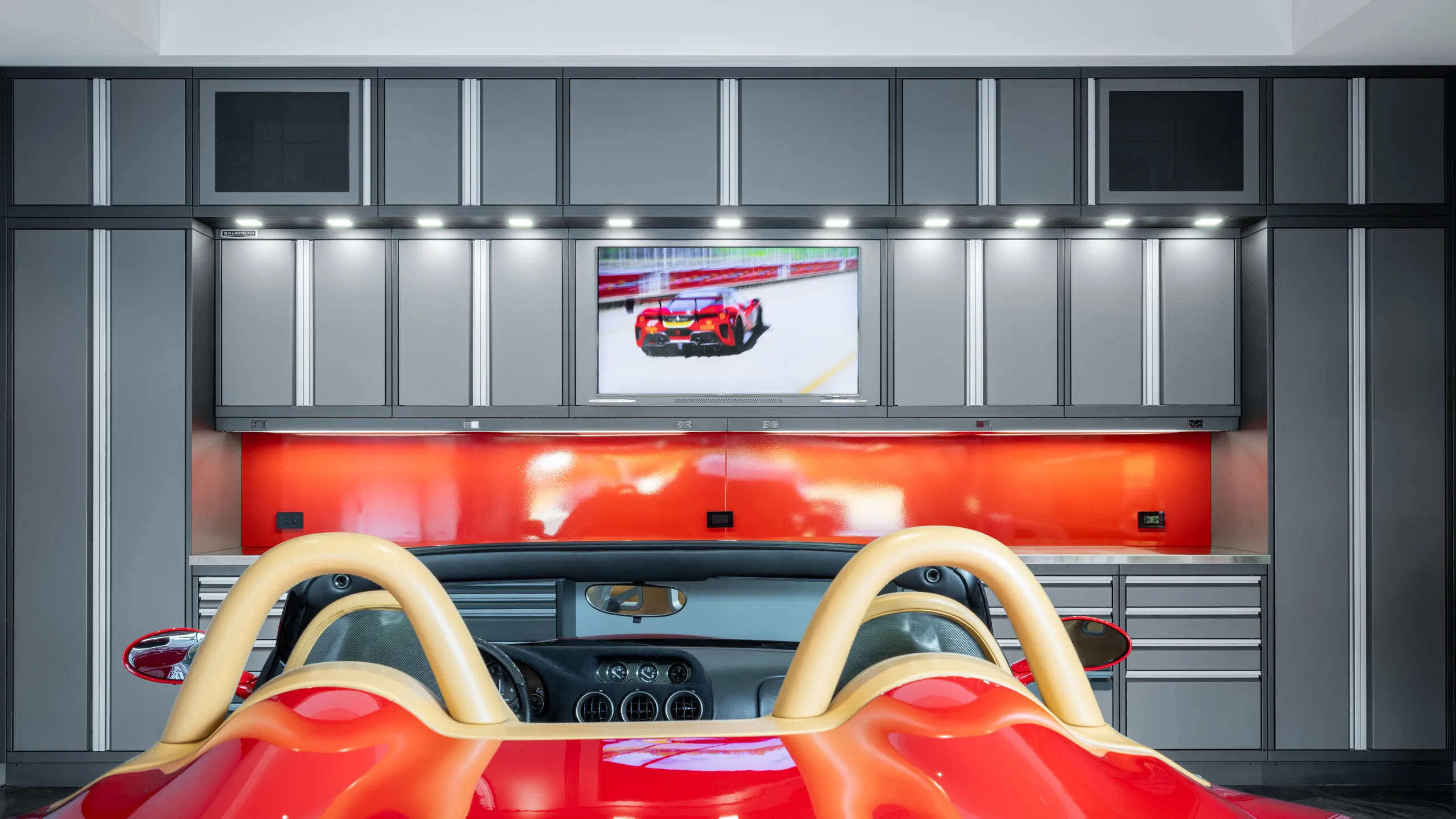 Luxury garage featuring custom Baldhead Cabinets with sleek gray finishes, integrated overhead lighting, bold red backsplash, and a high-end sports car parked in front of a wall-mounted TV displaying a racing scene.