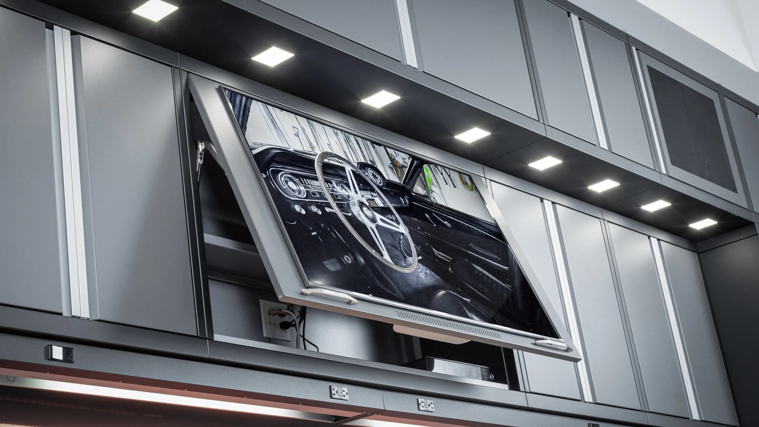 Custom garage cabinet with integrated flat-screen TV displaying a road view, high-end overhead LED lighting, and modern industrial storage by Baldhead Cabinets