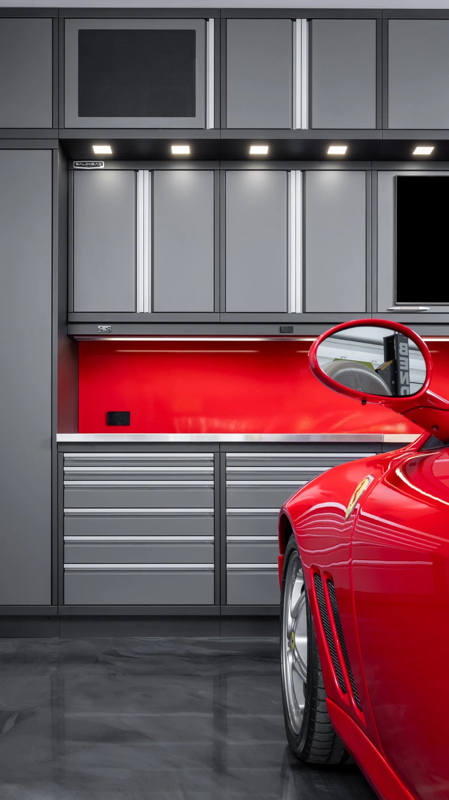 Modern garage with sleek gray Baldhead Cabinets, vibrant red backsplash, stainless steel worktop, and partial view of a red Ferrari, exemplifying luxury garage storage and design.
