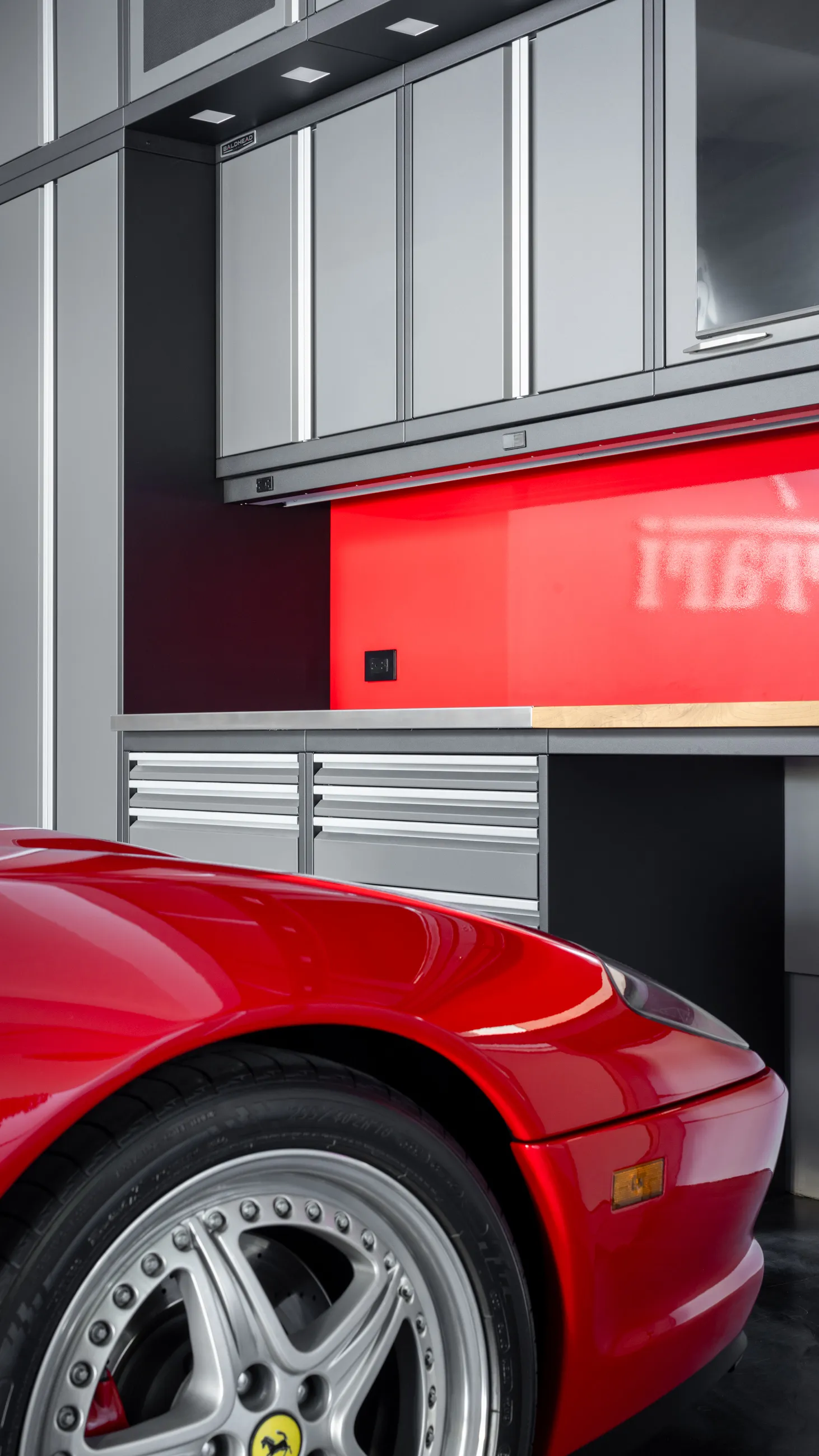 Luxury garage featuring Baldhead custom metal cabinets in matte gray, bold red backsplash, stainless steel work surface, and front view of a classic red Ferrari, emphasizing high-performance garage storage solutions.