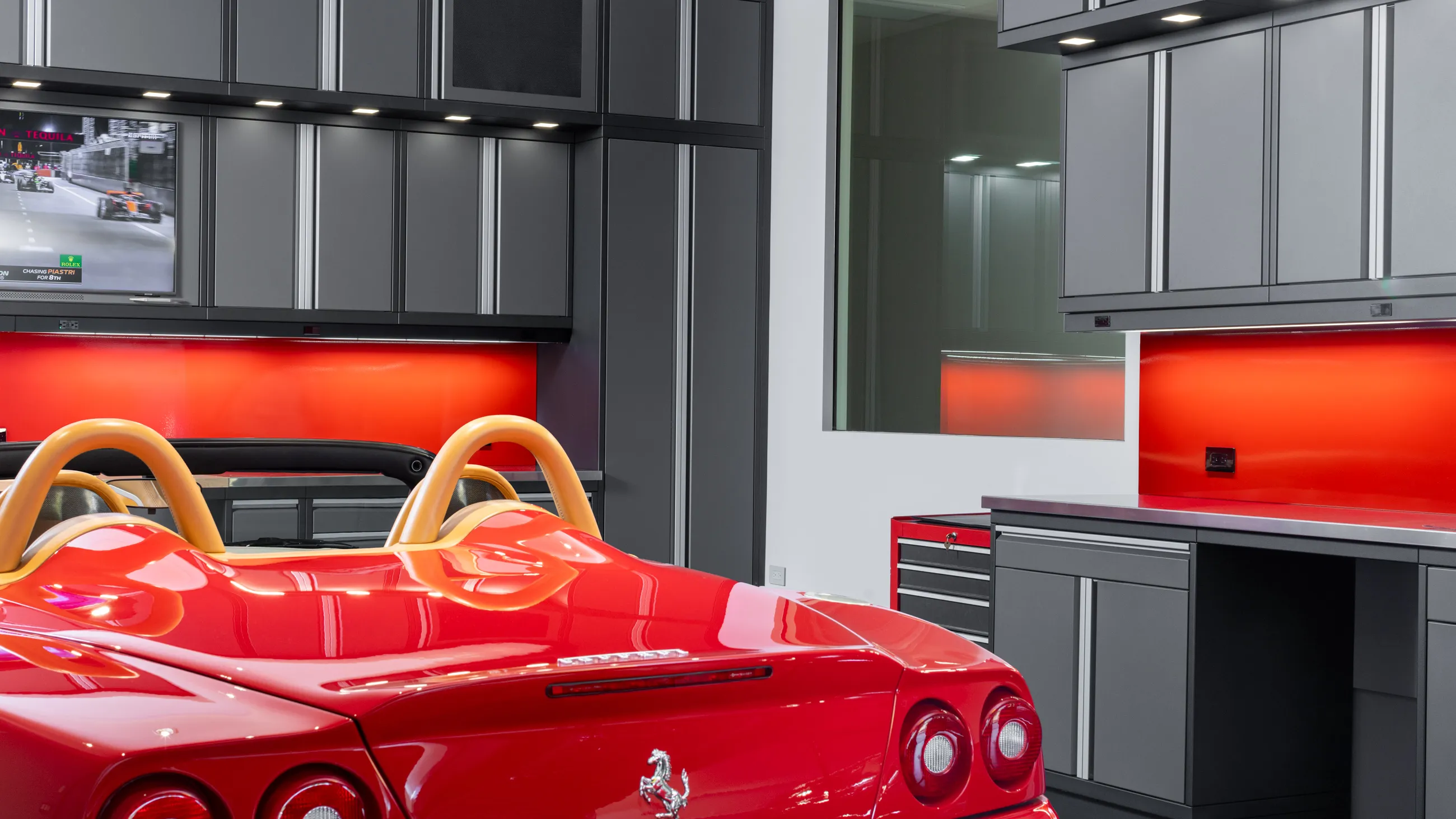 High-end garage with custom Baldhead Cabinets in sleek gray, bold red backsplash, built-in TV streaming motorsports, and a luxury red Ferrari convertible, highlighting premium garage organization and design.