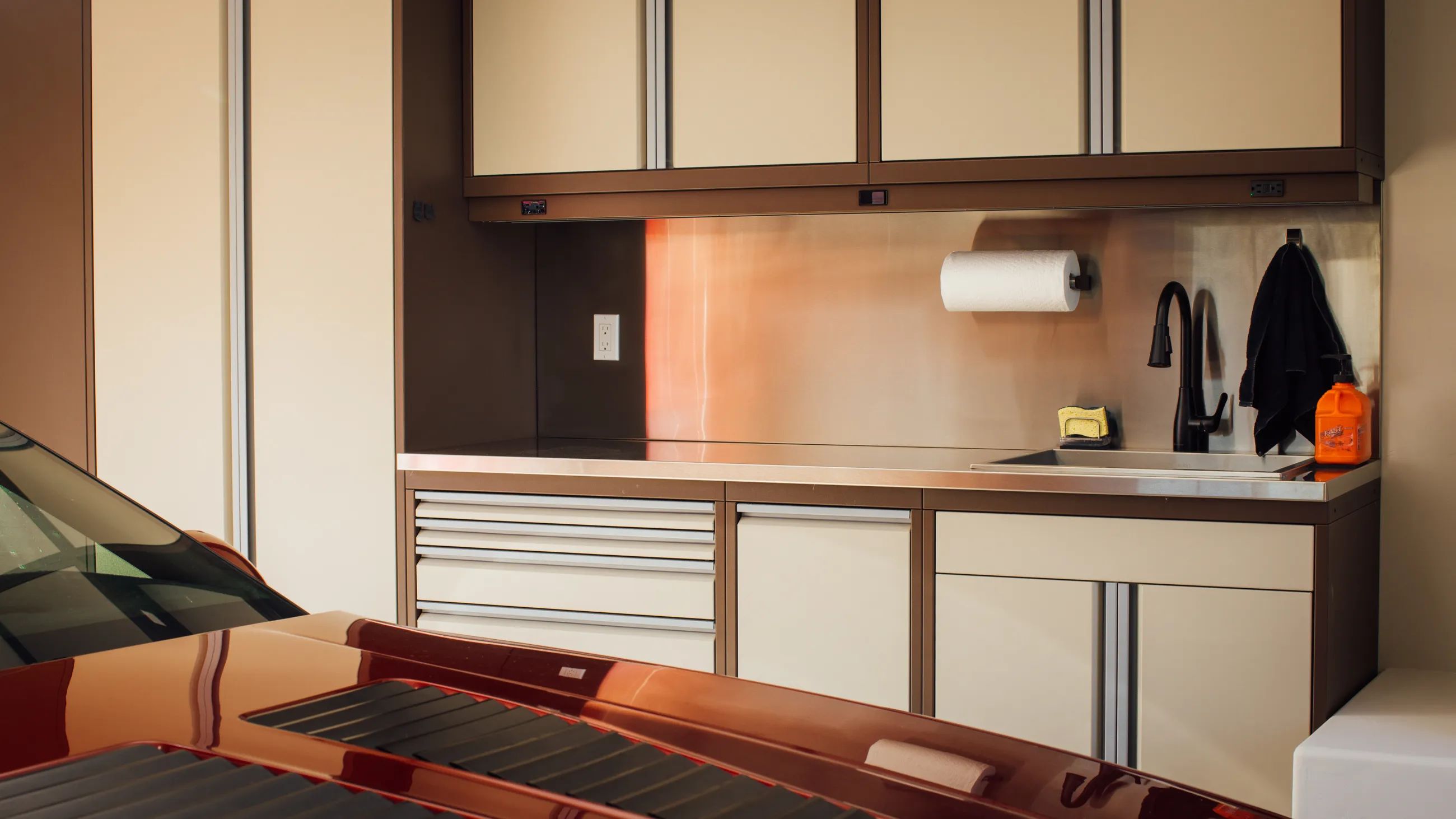 High-end garage station with custom Baldhead Cabinets in tan and brown finish, featuring stainless steel countertop and backsplash, black industrial faucet, under-cabinet lighting, and vintage sports car in the foreground.