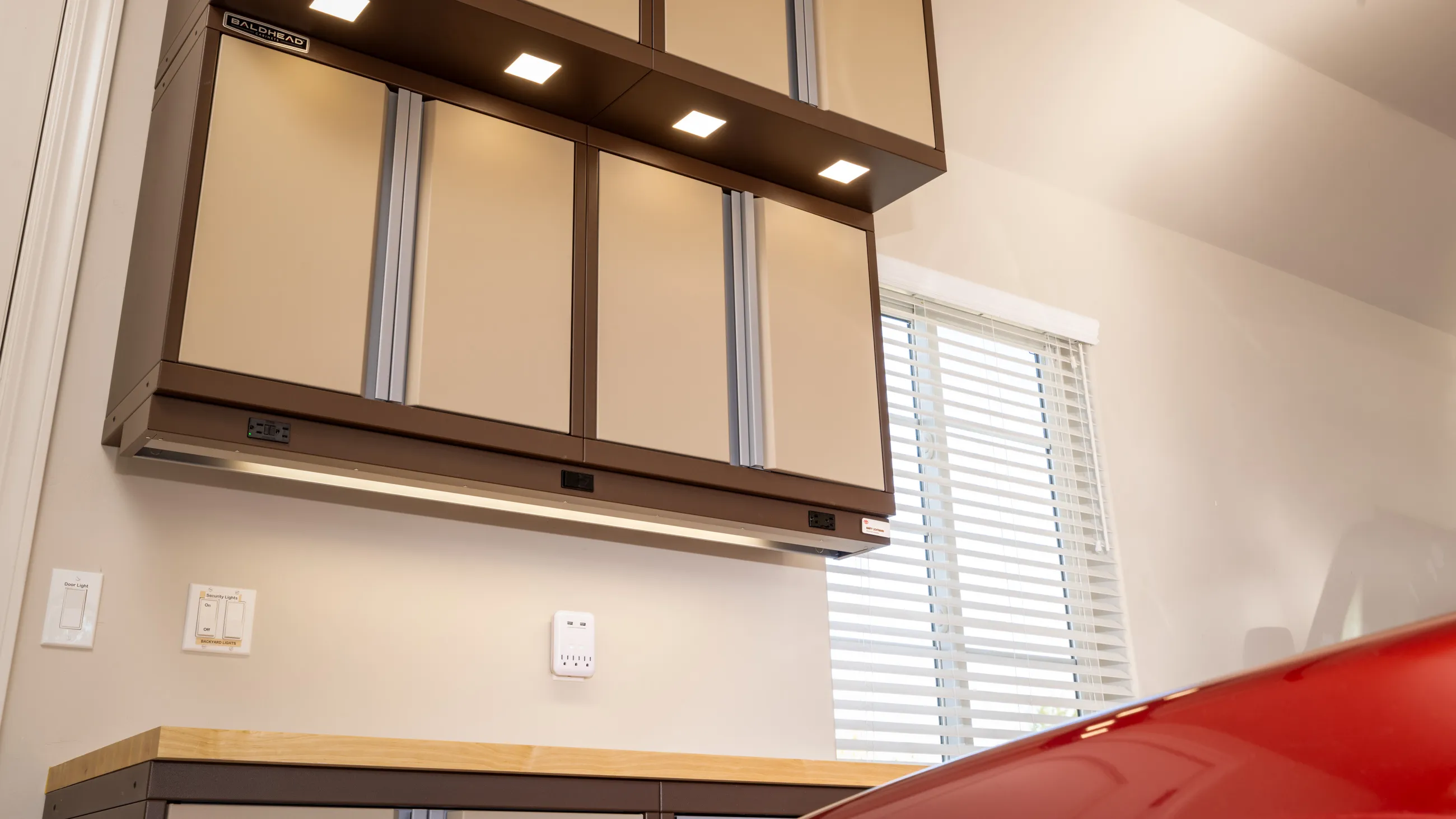 Garage workspace with custom Baldhead overhead cabinets in tan and brown finish, integrated LED lighting, butcher block countertop, and wall-mounted smart outlet, exemplifying modern and efficient garage storage design.