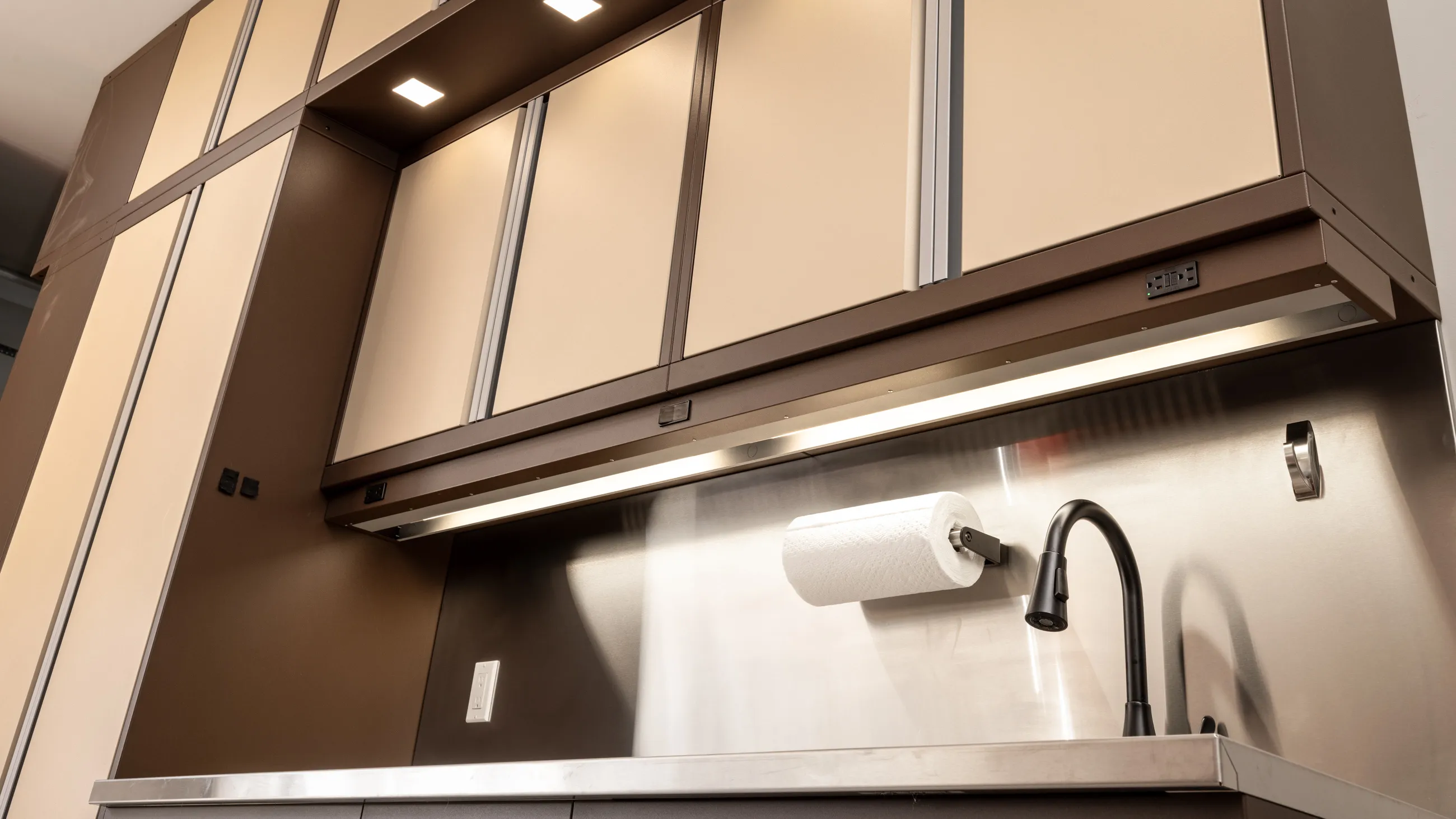 Custom Baldhead Cabinets in two-tone tan and brown finish with integrated lighting, stainless steel backsplash, under-cabinet outlets, industrial-style black faucet, and mounted paper towel holder for a high-functioning garage workspace.