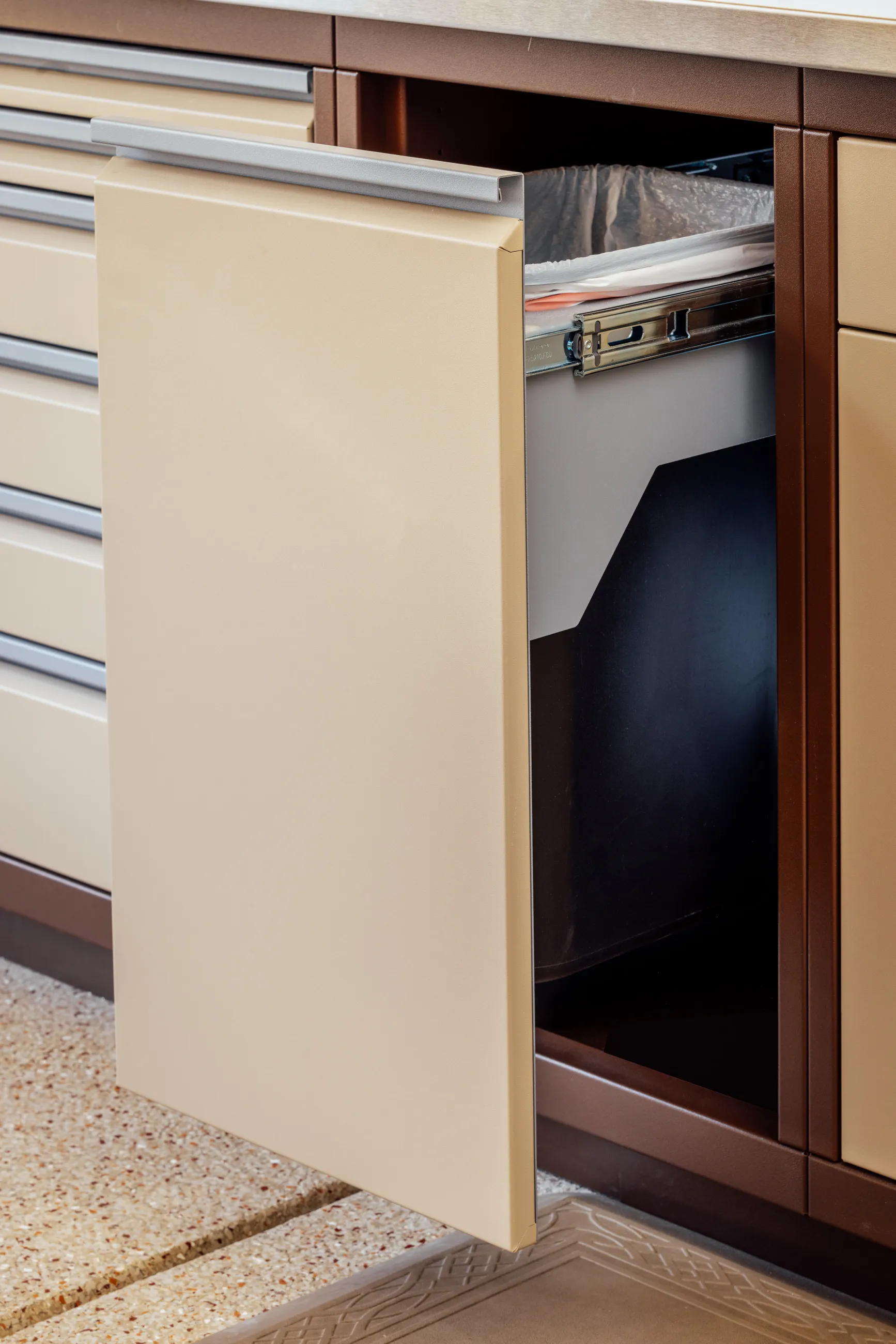 Custom Baldhead Cabinets with integrated pull-out trash bin in a two-tone tan and brown finish, designed for seamless functionality and clean aesthetics in luxury garage storage systems.