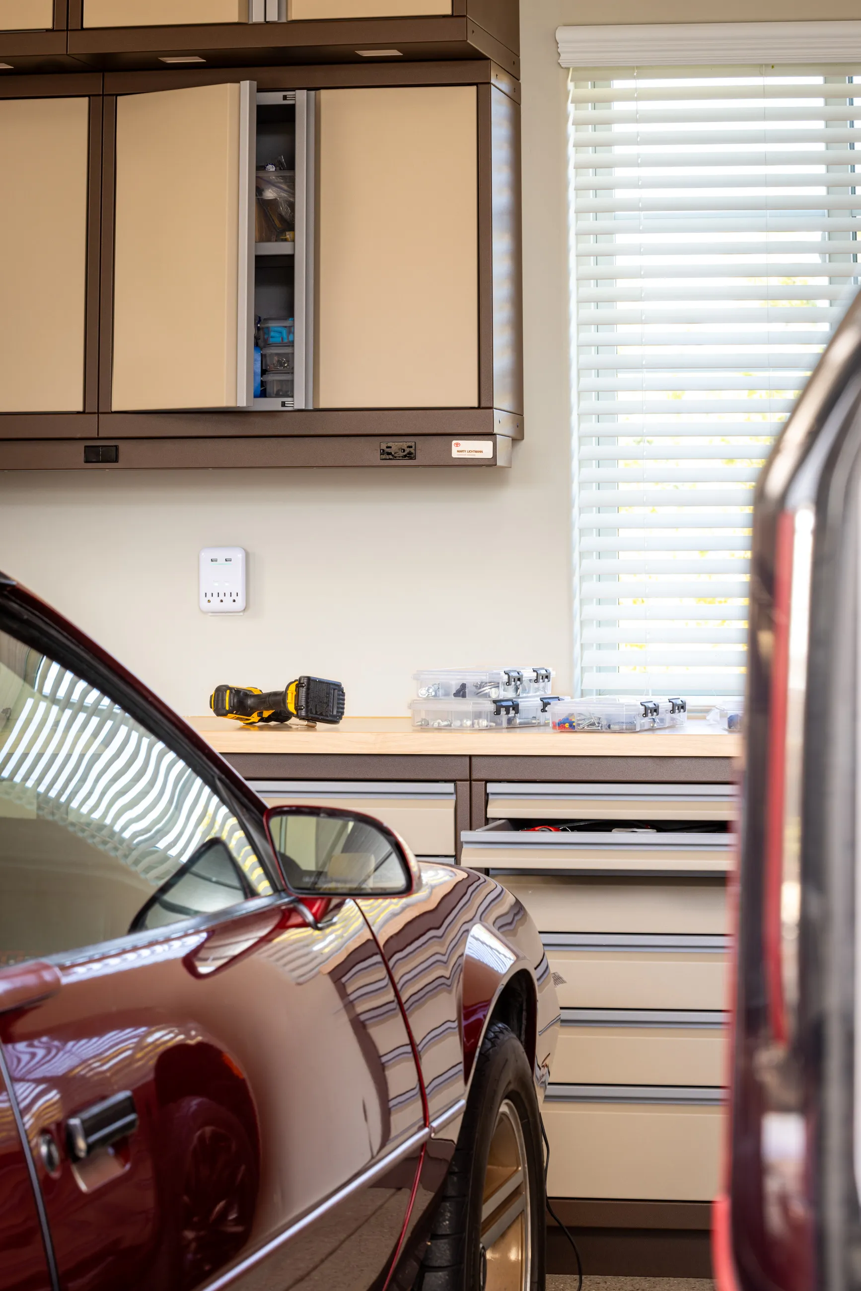 Organized garage workspace with Baldhead Cabinets in tan and brown, featuring overhead and base storage, a wood worktop with hardware kits and power tools, and a vintage red Camaro parked in front.