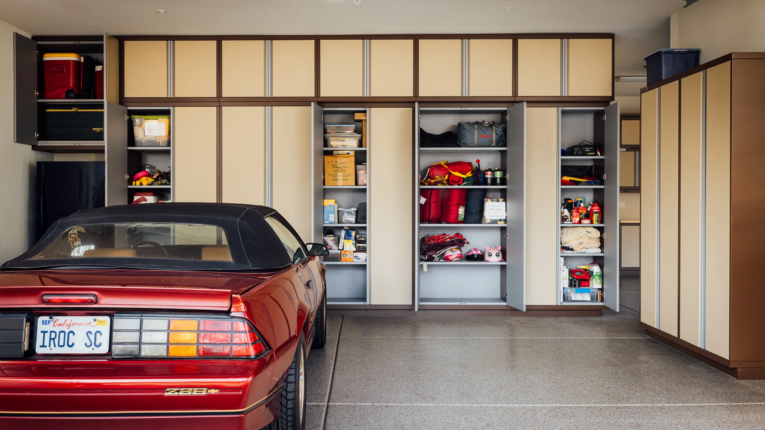 Custom garage storage cabinets by Baldhead Cabinets in a two-tone tan and bronze finish, shown with doors open to reveal organized shelves for tools, gear, and supplies—perfect for maximizing storage in high-end garage setups alongside classic cars.