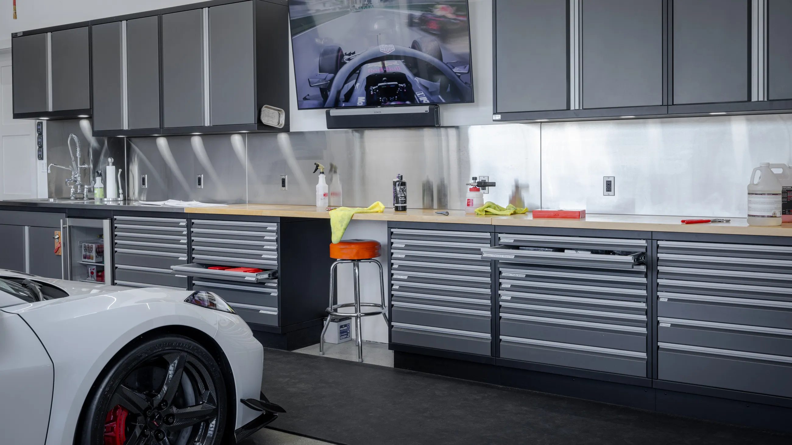 Luxury garage workstation with Baldhead Cabinets' custom-built modular steel cabinetry, featuring full-extension drawers, overhead storage, and stainless steel countertops. Equipped with detailing products, high-end fixtures, and a wall-mounted racing monitor for an elevated workshop experience. Ideal for automotive enthusiasts seeking premium garage organization.