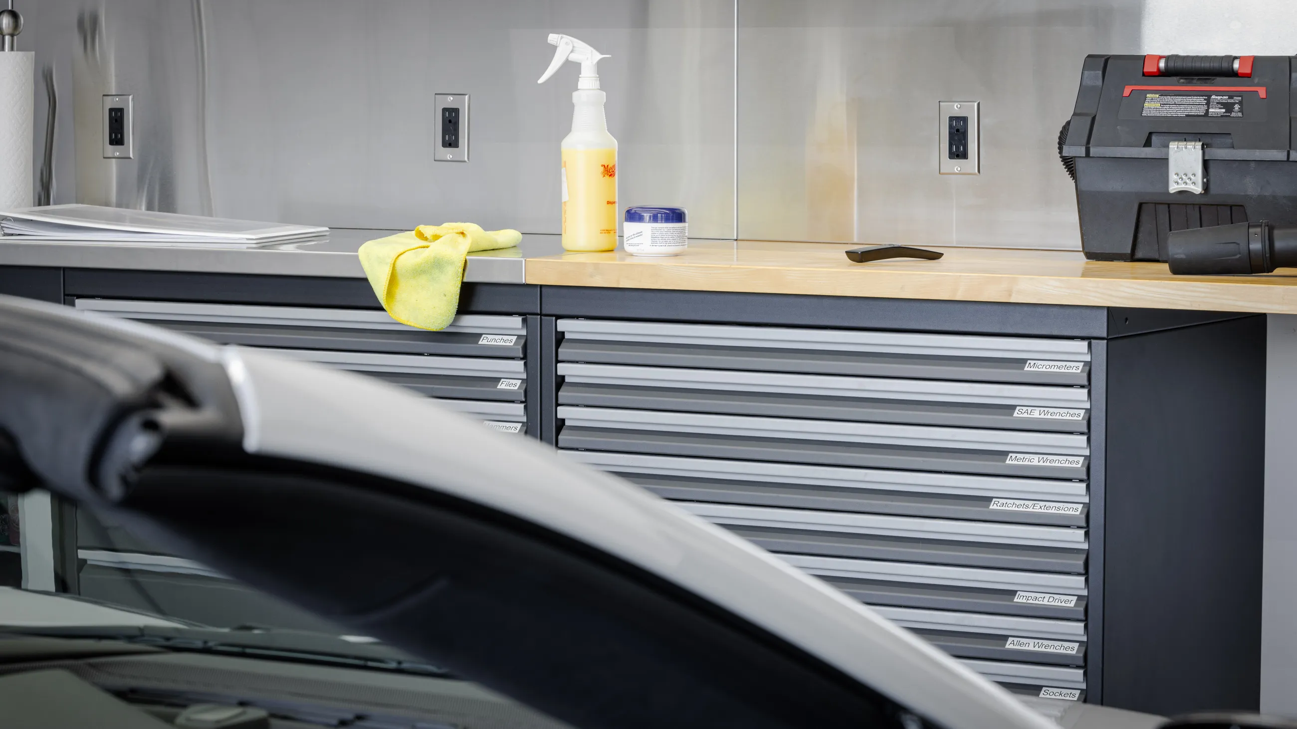 Premium garage cabinet workstation featuring Baldhead Cabinets’ durable steel drawers, labeled for organized tool storage including micrometers, SAE and metric wrenches, and sockets. Clean stainless steel backsplash, butcher block countertop, and detailing supplies highlight a high-end automotive workshop setup.