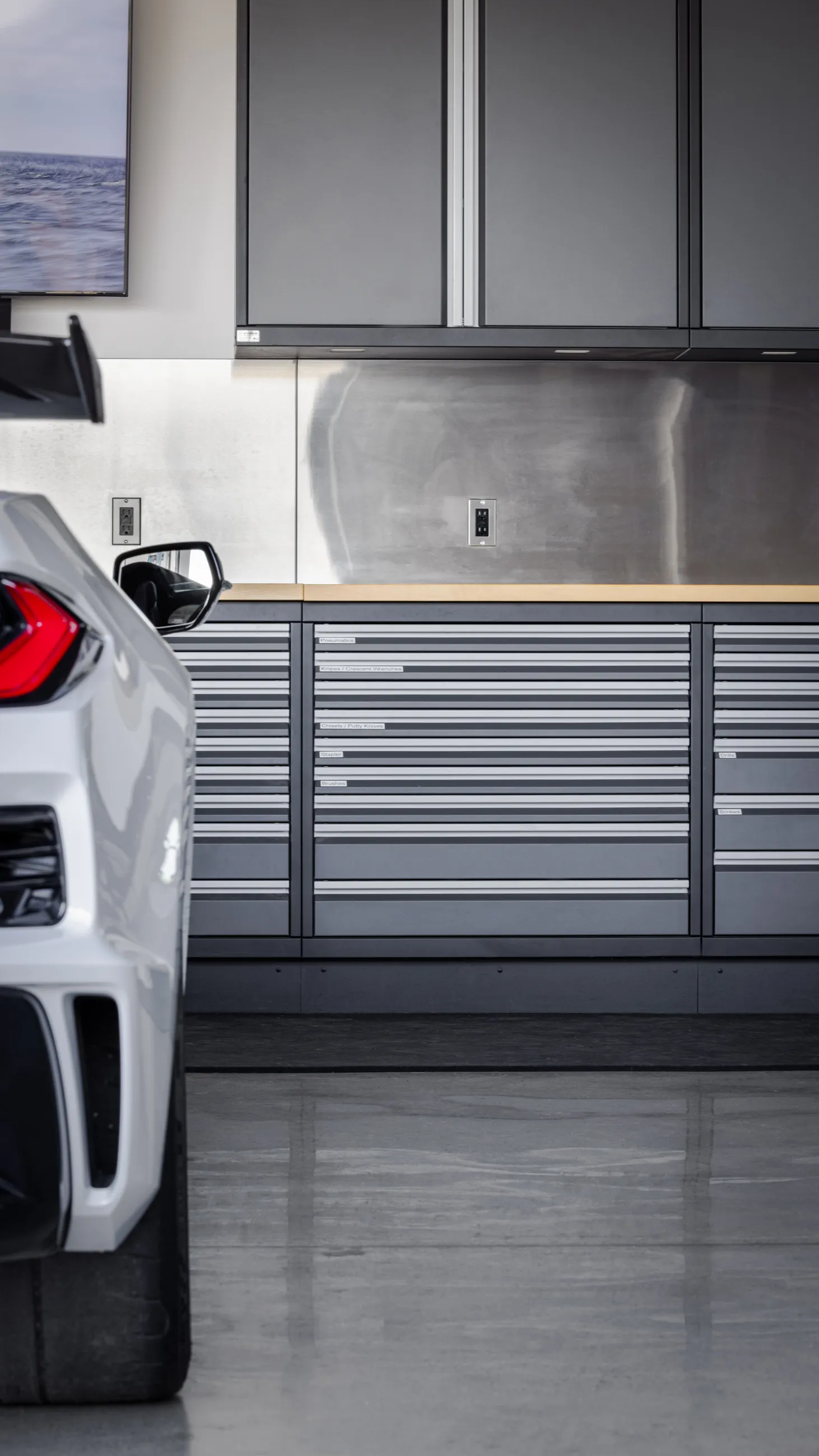Modern luxury garage setup featuring Baldhead Cabinets’ sleek steel drawer systems and overhead cabinets, paired with a stainless steel backsplash and maple countertop. A white performance sports car adds a high-end touch, highlighting the brand’s commitment to precision-crafted garage storage for automotive enthusiasts.