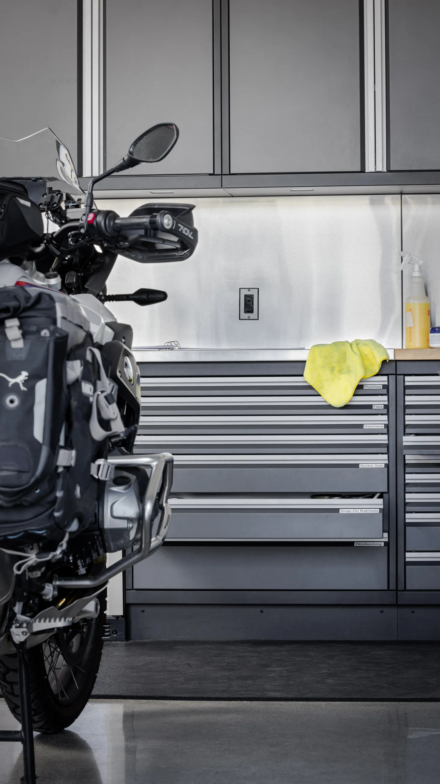 Adventure-ready garage featuring Baldhead Cabinets’ precision-crafted steel storage drawers labeled for punches, files, hammers, and Snap-On ratchets. Mounted beneath sleek overhead cabinets and stainless backsplash, this organized workstation supports motorcycle maintenance and premium tool organization. Ideal for riders and gearheads seeking durable garage solutions.