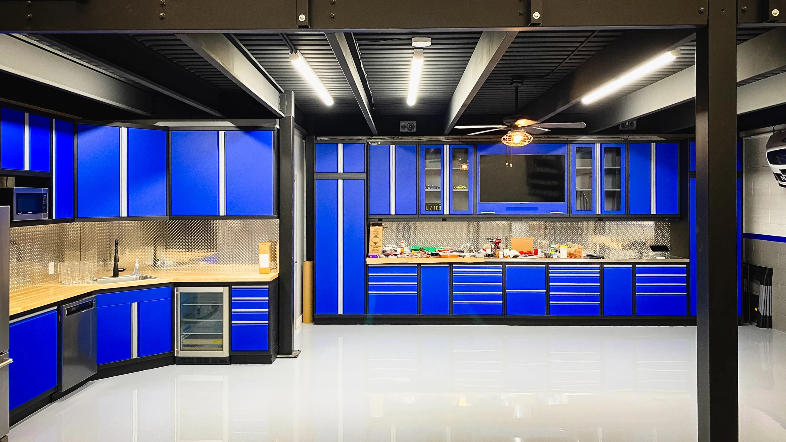 Custom garage featuring vibrant blue Baldhead Cabinets with sleek aluminum trim and natural wood countertops. Diamond plate backsplash adds industrial flair, while built-in appliances and under-cabinet lighting enhance functionality. Glossy epoxy floor reflects premium craftsmanship and modern design, showcasing Baldhead Cabinets’ high-end garage organization and cabinetry solutions.