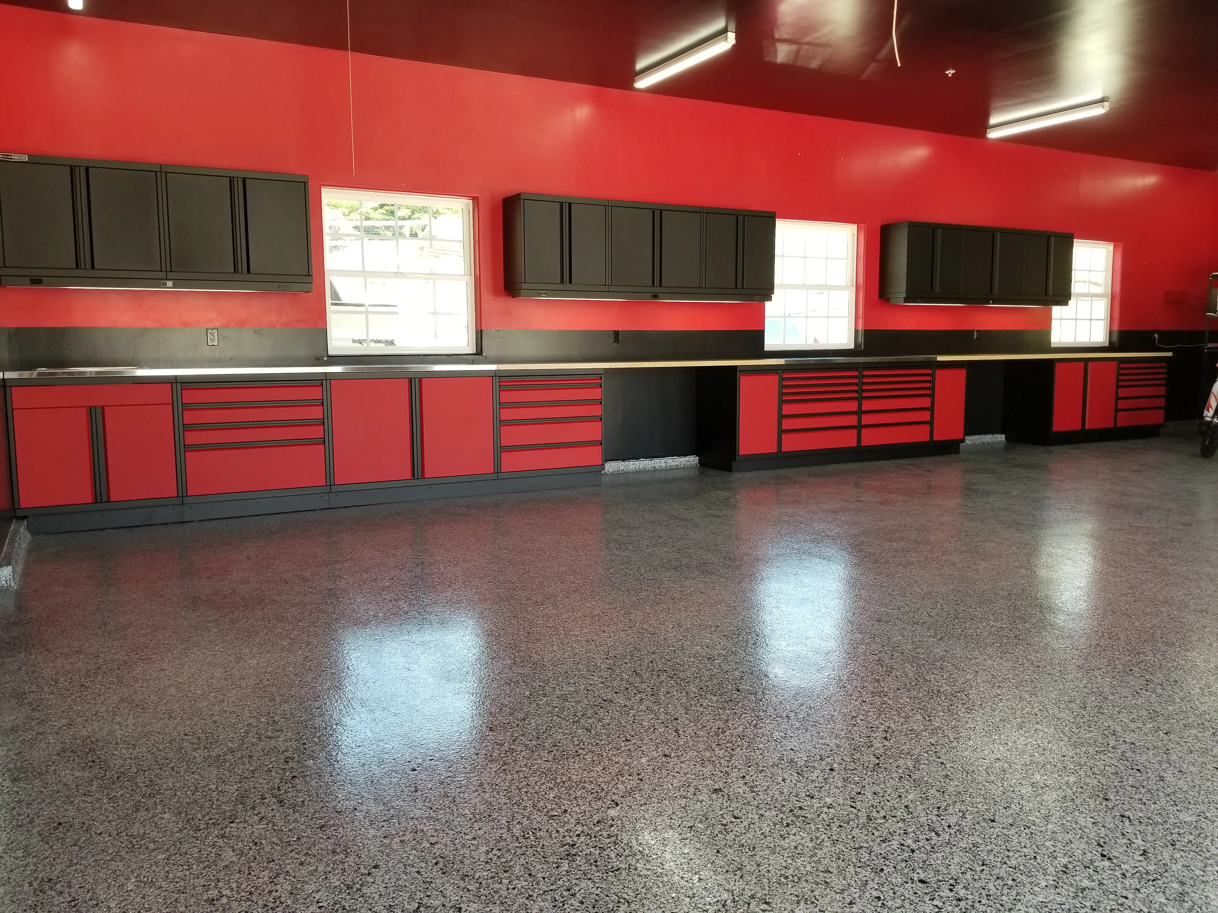 Custom high-performance garage storage system featuring bold red and black Baldhead Cabinets with sleek aluminum trim, ample drawer space, upper wall-mounted cabinets, and a polished epoxy floor—designed for serious workspace efficiency and striking visual appeal.