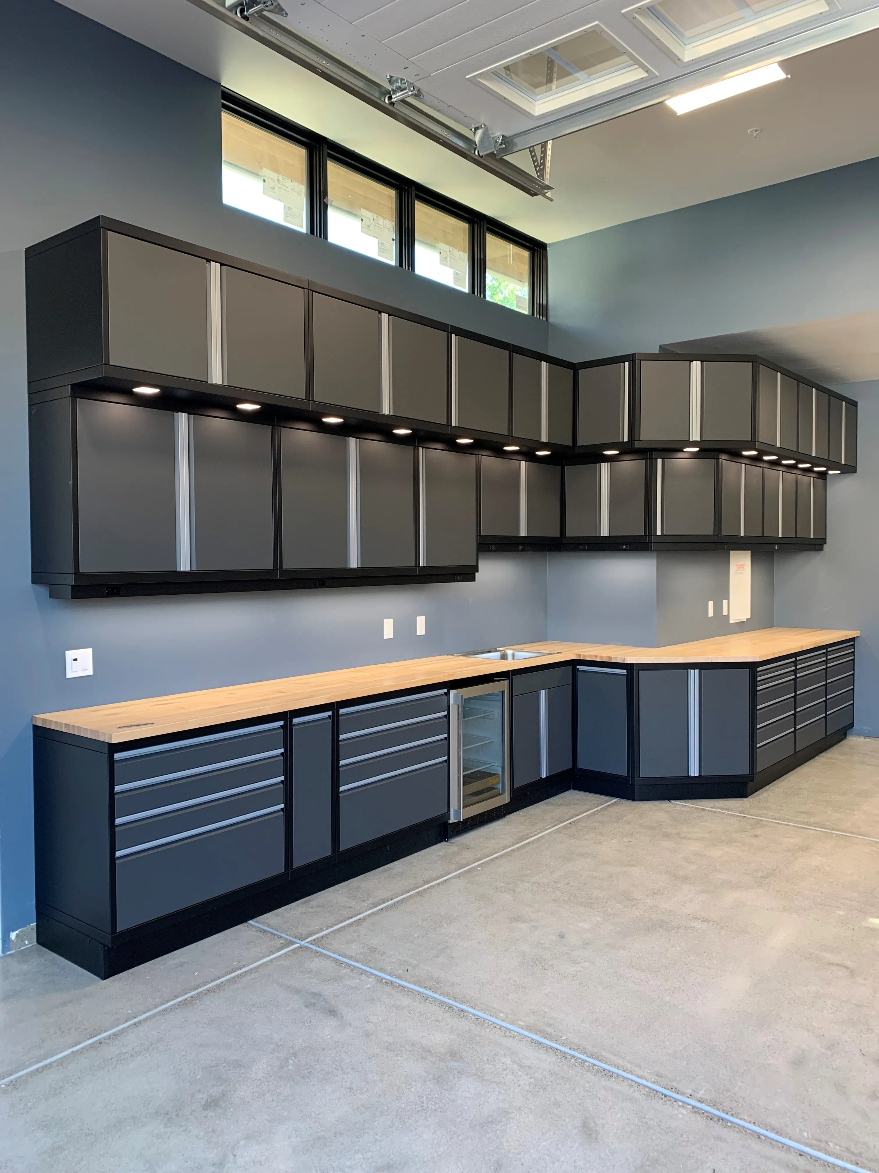 L-shaped custom garage cabinets by Baldhead Cabinets in matte charcoal finish with silver trim, featuring upper and lower storage, under-cabinet LED lighting, butcher block countertops, integrated sink, and beverage cooler in a high-end workshop environment.