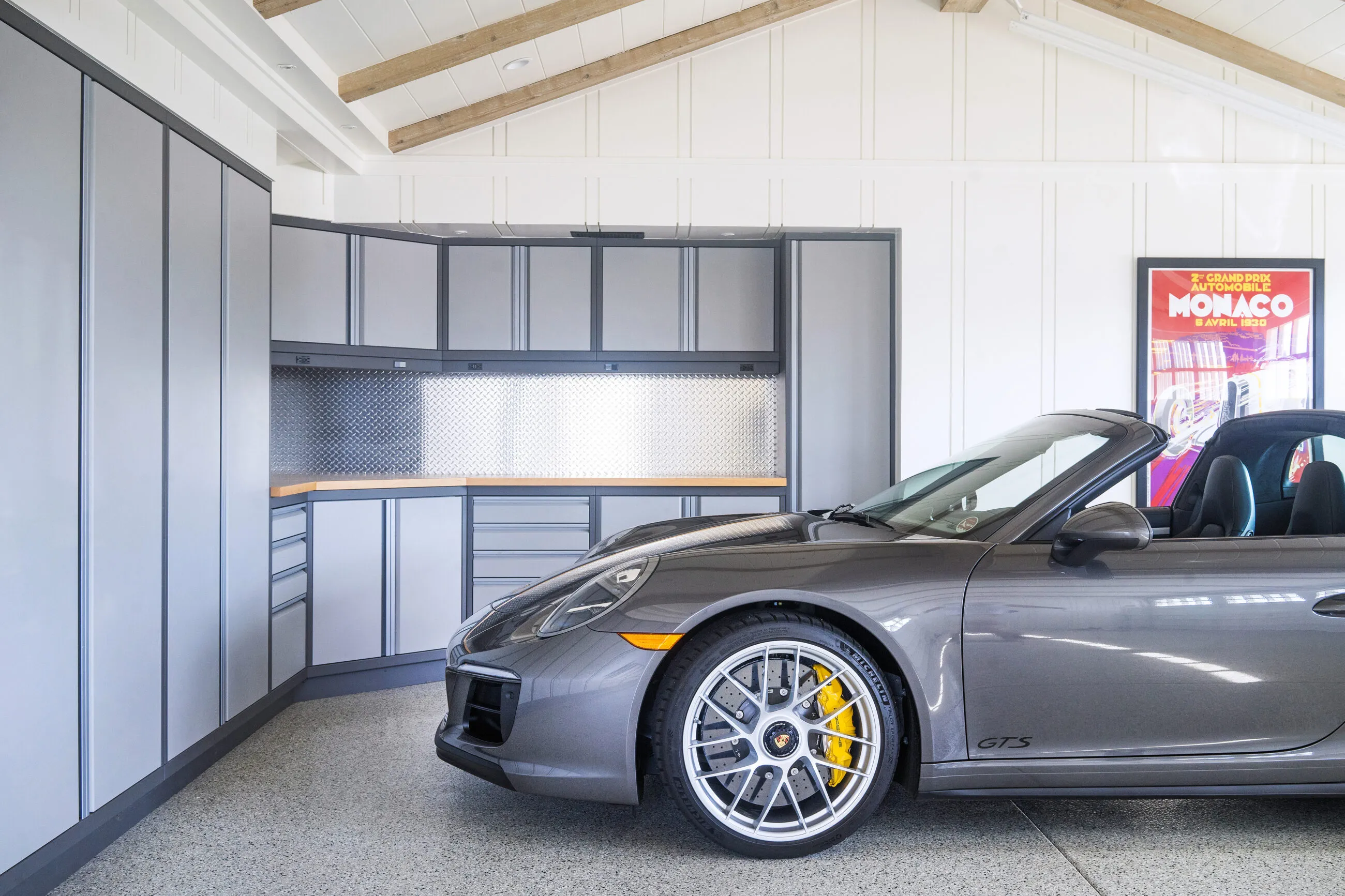 Luxury garage featuring custom Baldhead Cabinets in a sleek gray finish with vertical trim accents. Diamond plate aluminum backsplash adds an industrial touch, paired with a warm wood countertop. Showcased with a Porsche 911 GTS, this premium garage setup blends performance and style, ideal for high-end automotive storage and workspace solutions.
