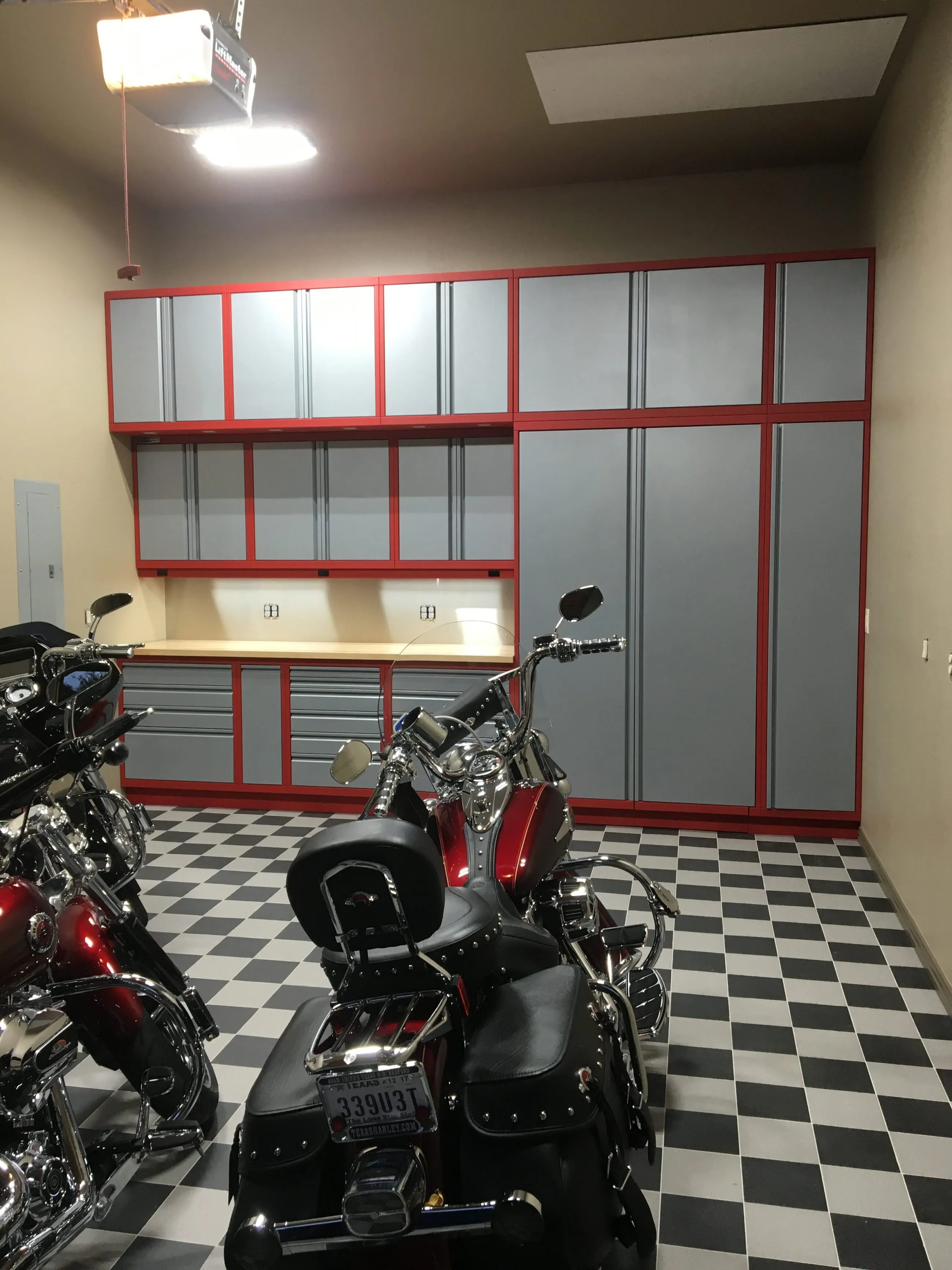 Custom red and gray Baldhead Cabinets in a motorcycle garage with checkered tile flooring, integrated upper and lower storage, butcher block countertops, and multiple Harley-Davidson bikes showcasing high-end garage organization and style.