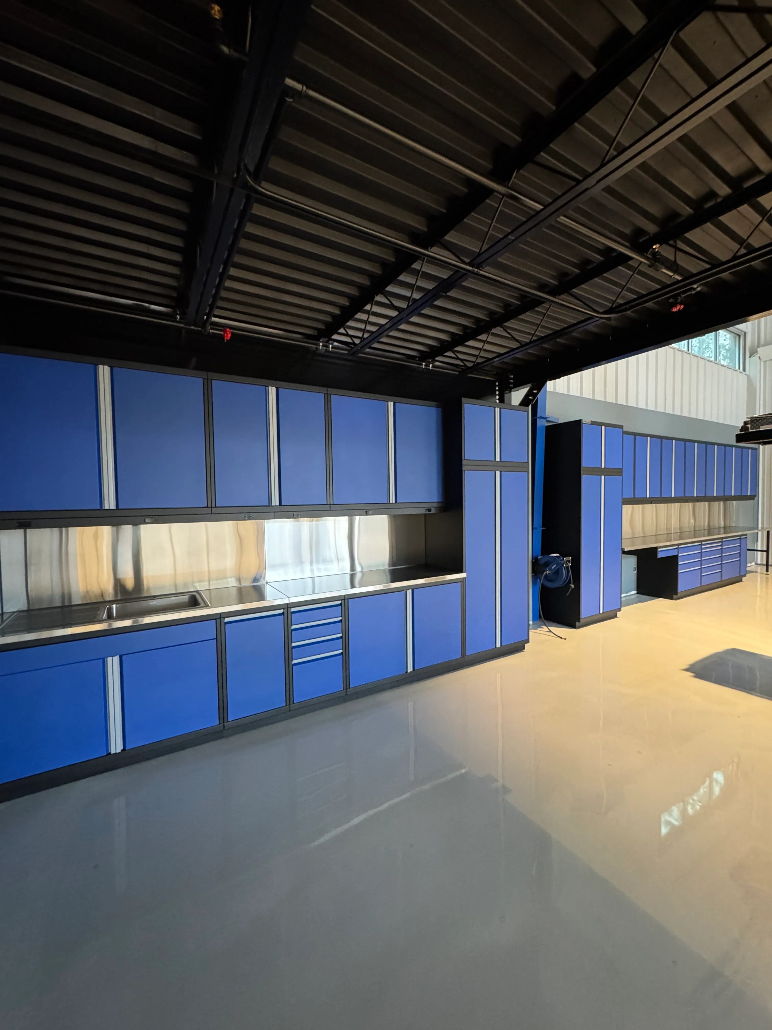 Bold blue custom garage cabinets by Baldhead Cabinets, featuring powder-coated steel doors, stainless steel countertops with integrated sink, wall-mounted hose reel, and a clean epoxy-coated floor in a high-end automotive workspace.