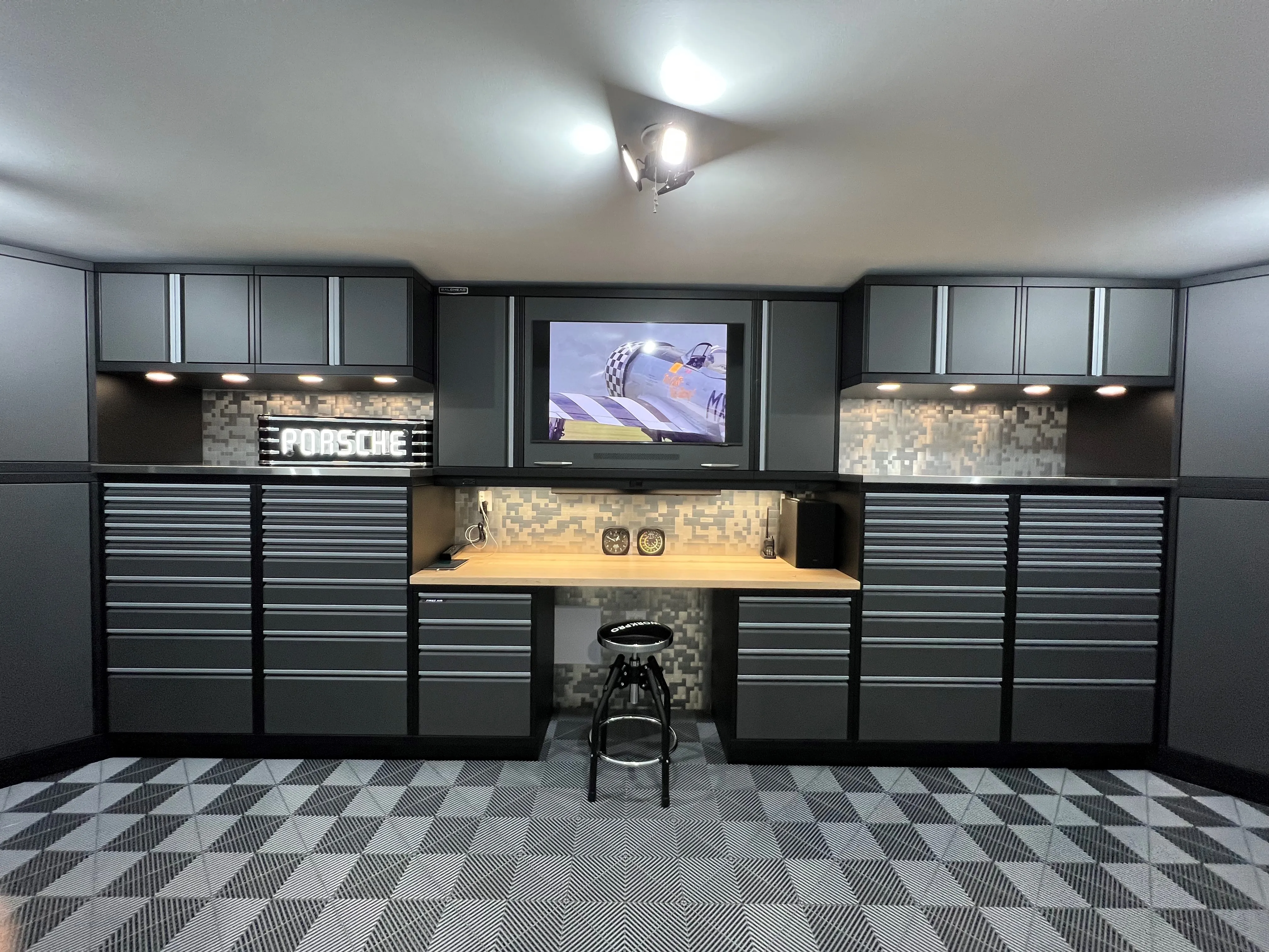 High-performance garage workstation by Baldhead Cabinets featuring matte black powder-coated cabinets, integrated TV, LED task lighting, mosaic tile backsplash, Porsche signage, and custom modular flooring in a motorsport-inspired luxury workspace.