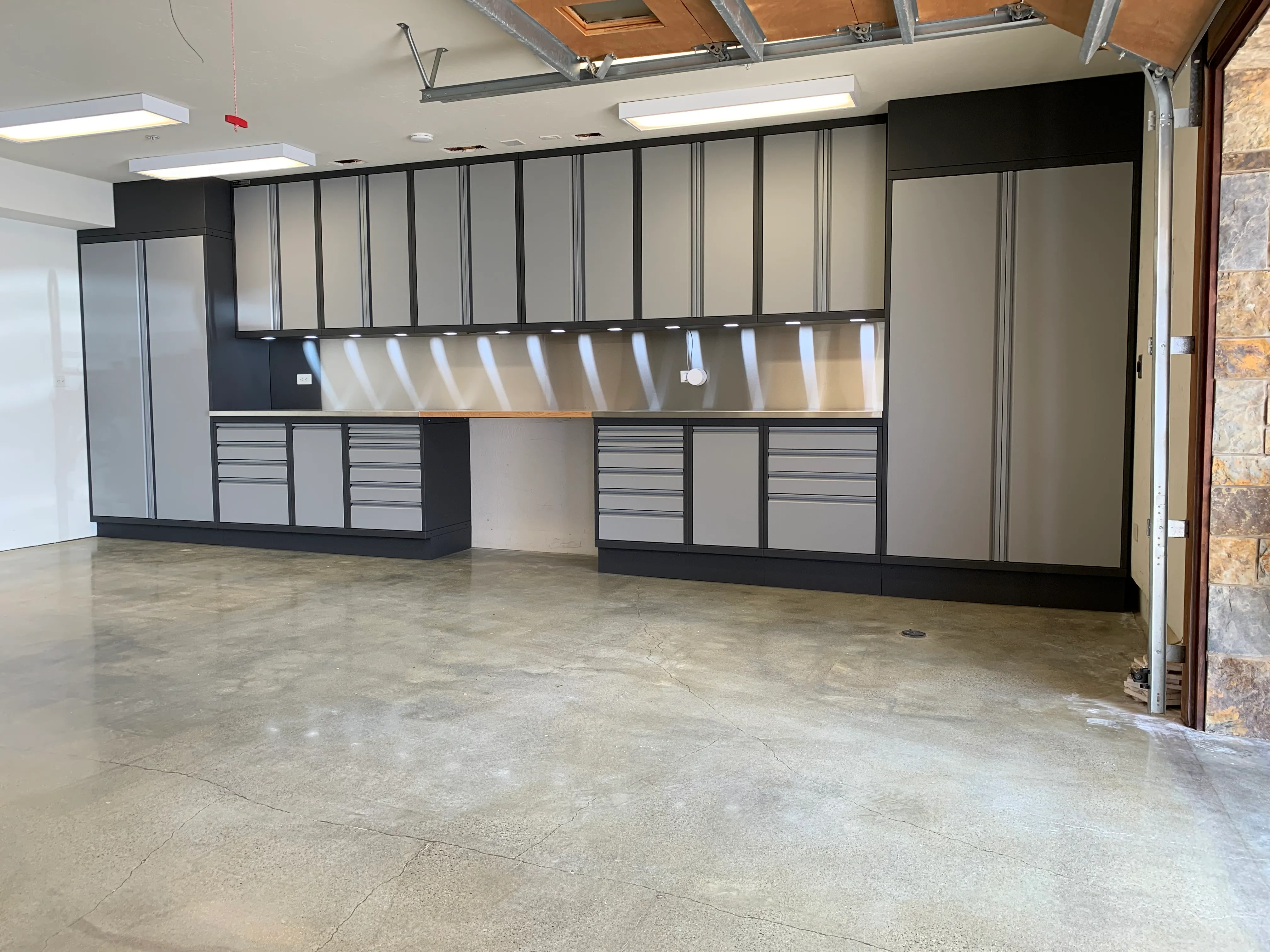 Custom two-tone garage cabinets by Baldhead Cabinets featuring matte gray and black powder-coated finishes, upper and lower storage, integrated LED task lighting, stainless steel backsplash, and polished concrete flooring in a modern residential garage.