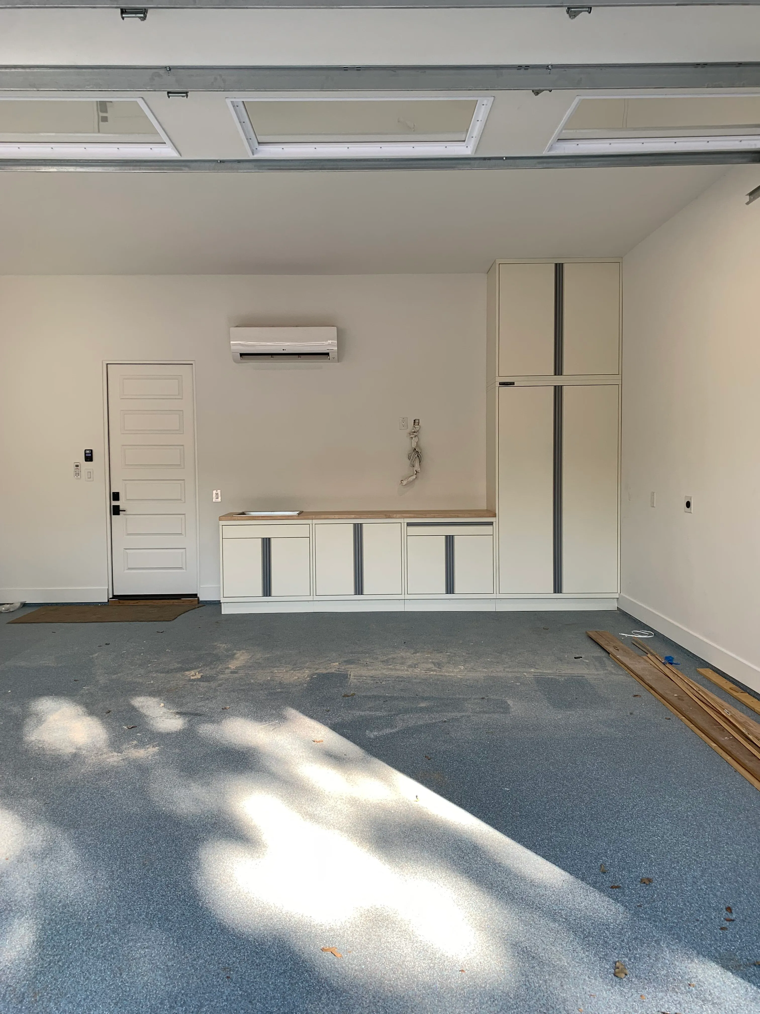Minimalist garage setup by Baldhead Cabinets featuring cream-colored powder-coated cabinetry with gray accents, integrated tall storage, butcher block countertop, and light blue epoxy flooring in a clean, climate-controlled residential garage.
