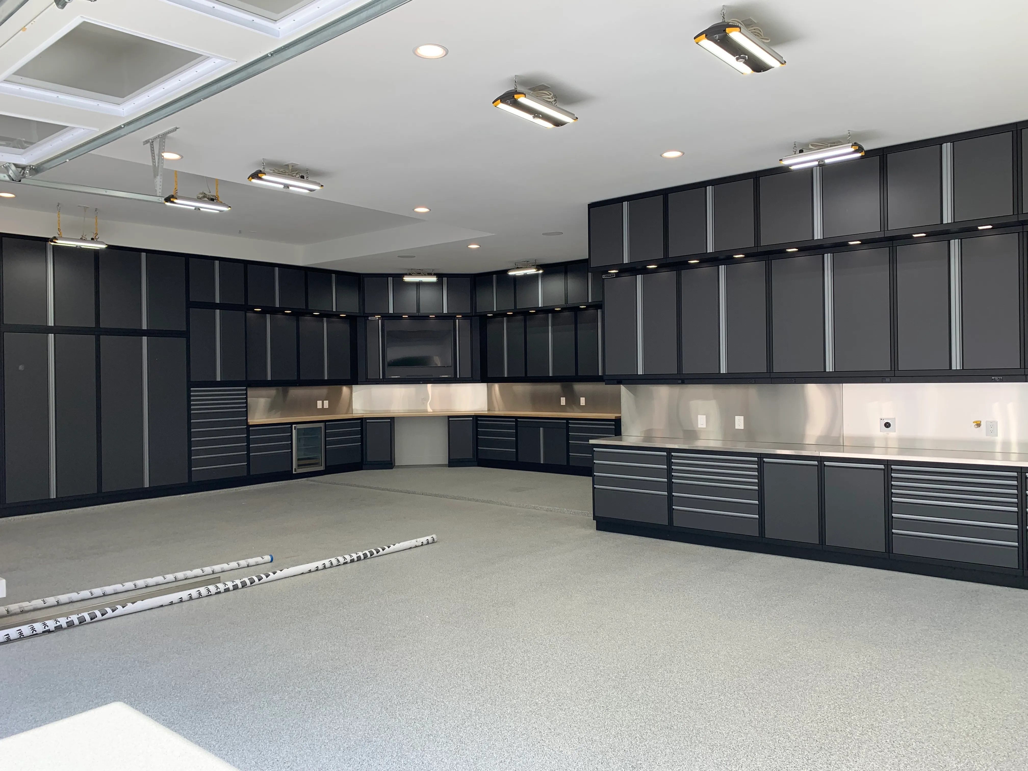 Spacious custom garage featuring Baldhead Cabinets’ matte black and gray aluminum storage system with full-height wall cabinets, under-cabinet LED lighting, stainless steel countertops and backsplash, integrated beverage fridge, and TV cutout—perfect for high-end garage organization and workspace solutions.