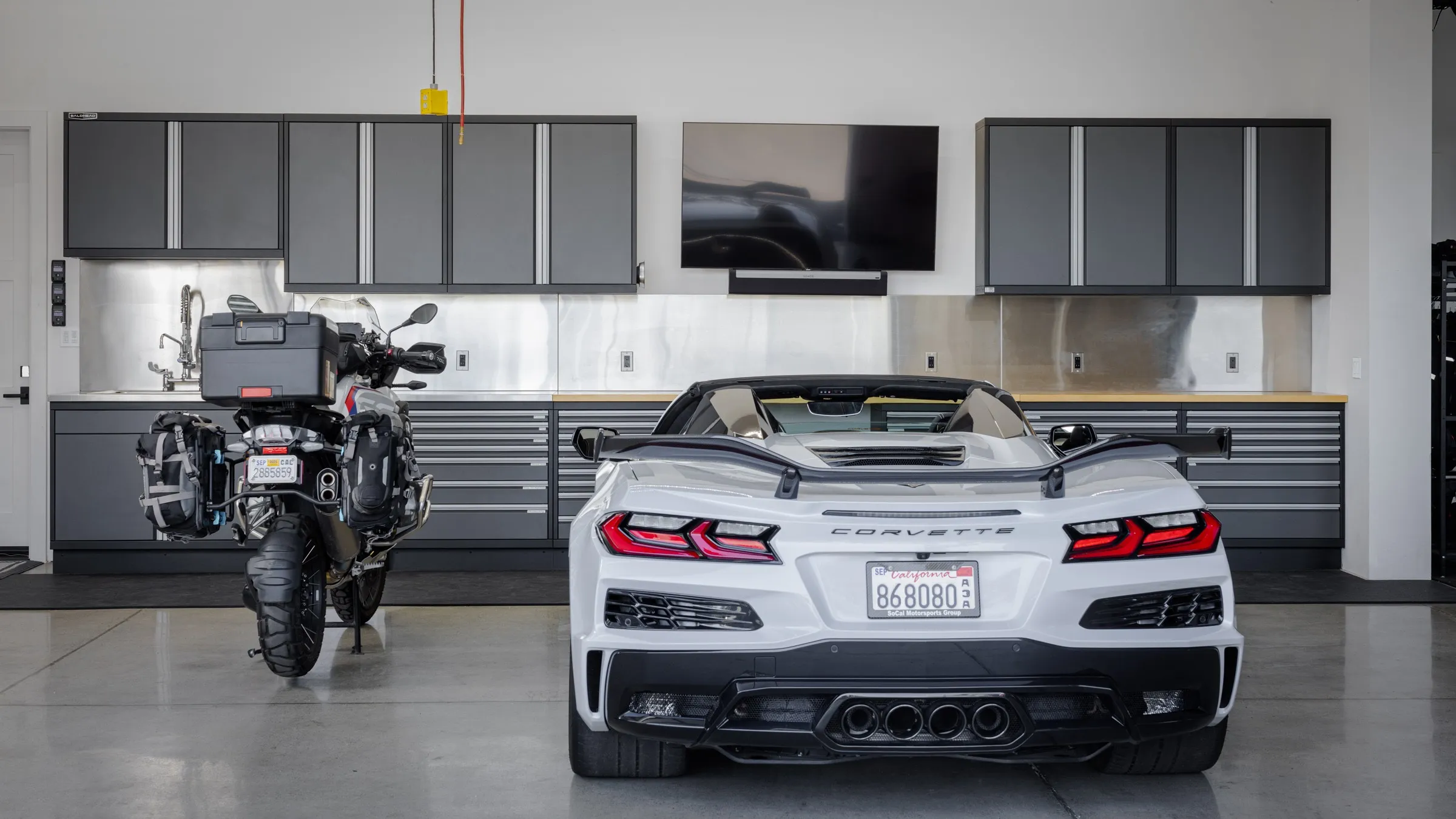 Showcase garage featuring Baldhead Cabinets’ premium custom metal cabinetry, sleek upper storage, and stainless steel backsplash. A white Corvette convertible and adventure motorcycle complete the luxury automotive setup, highlighting high-performance garage organization for car collectors and enthusiasts.