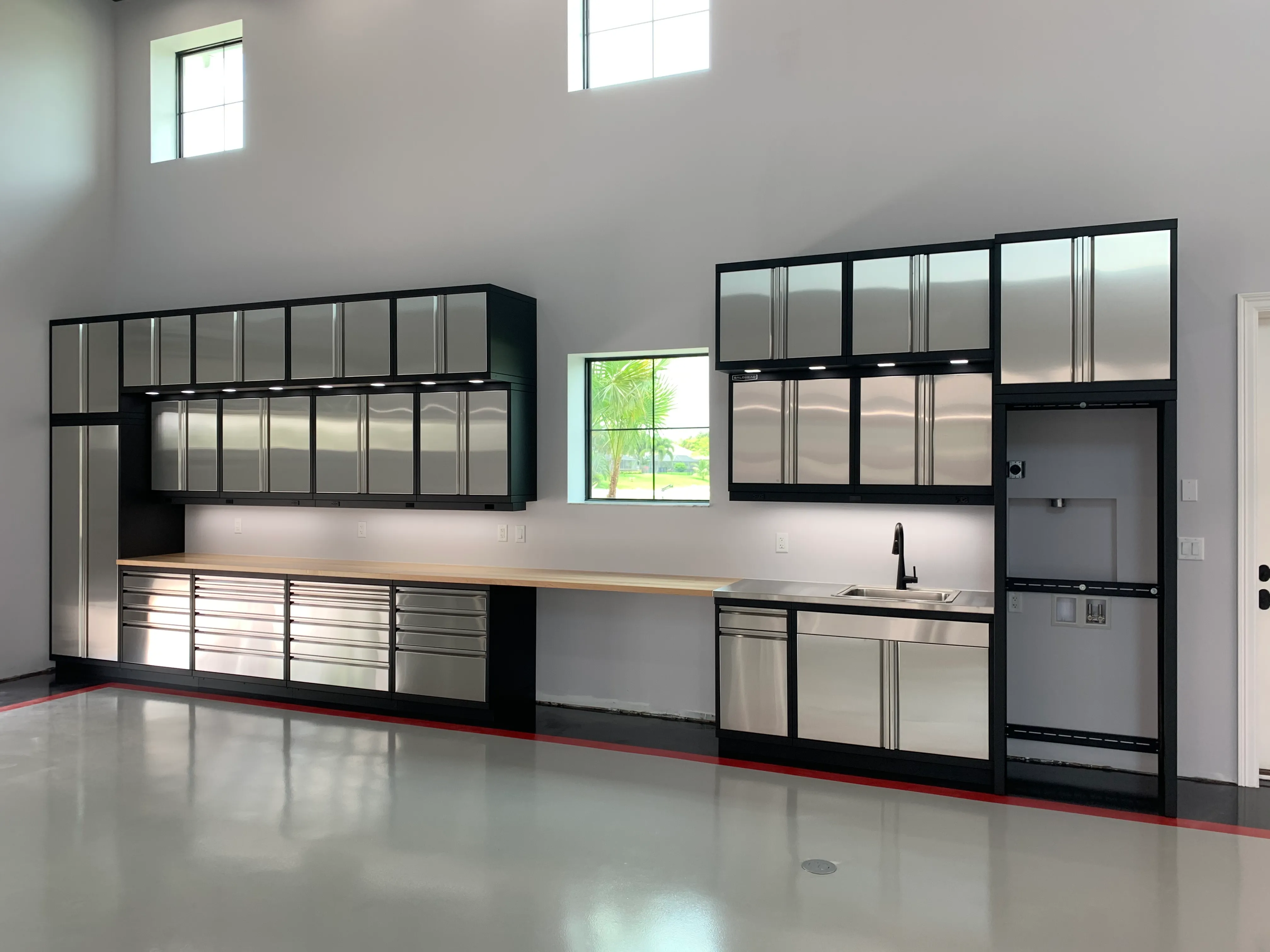 High-end stainless steel garage cabinets by Baldhead Cabinets featuring upper and lower modular storage, integrated sink, butcher block and stainless countertops, appliance-ready space, and LED task lighting in a luxury residential garage.