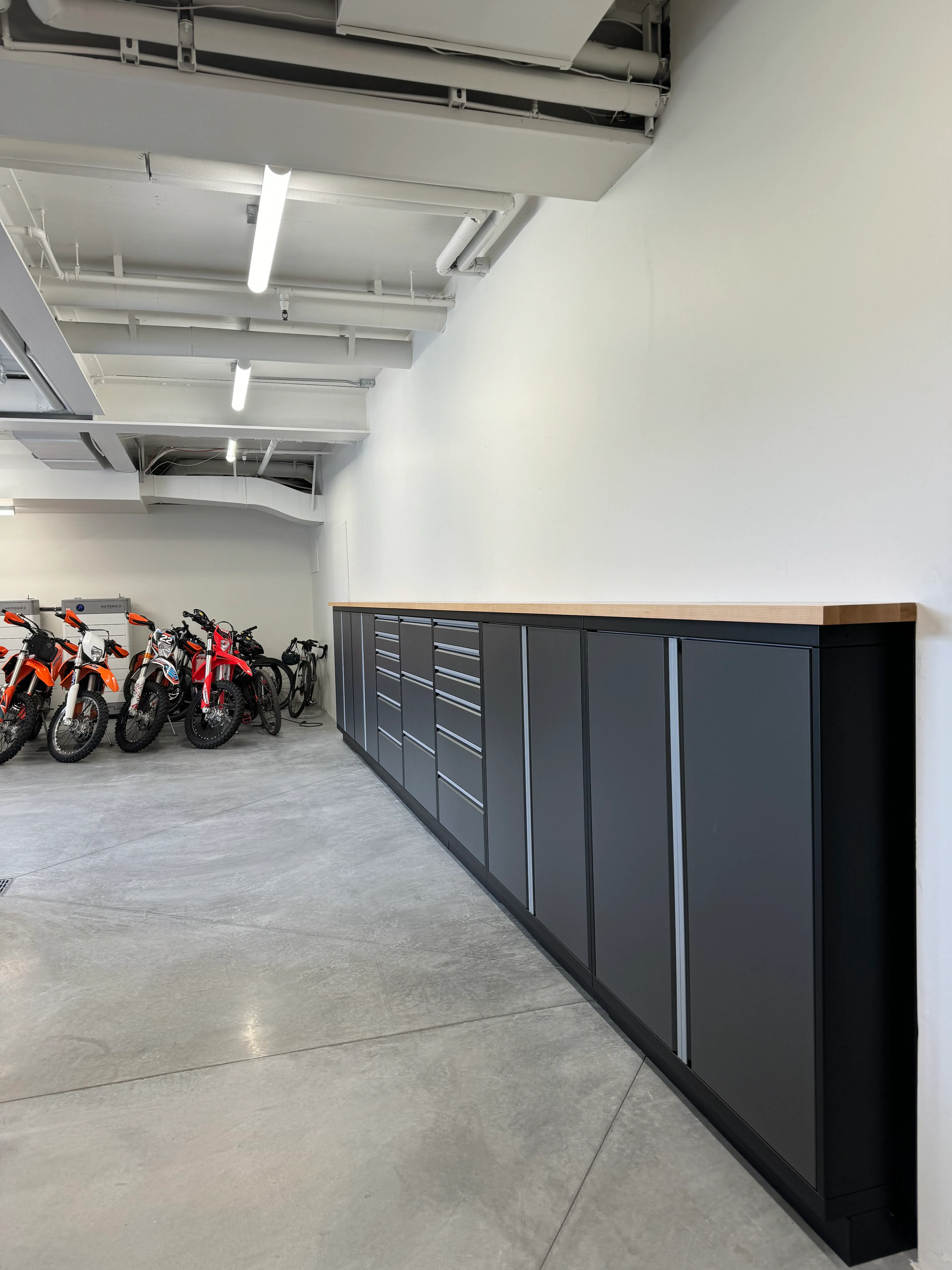 Streamlined matte gray Baldhead Cabinets with integrated drawers and butcher block countertop, installed in a polished concrete garage with dirt bikes and bicycles, offering rugged, high-performance storage for motorsport enthusiasts.