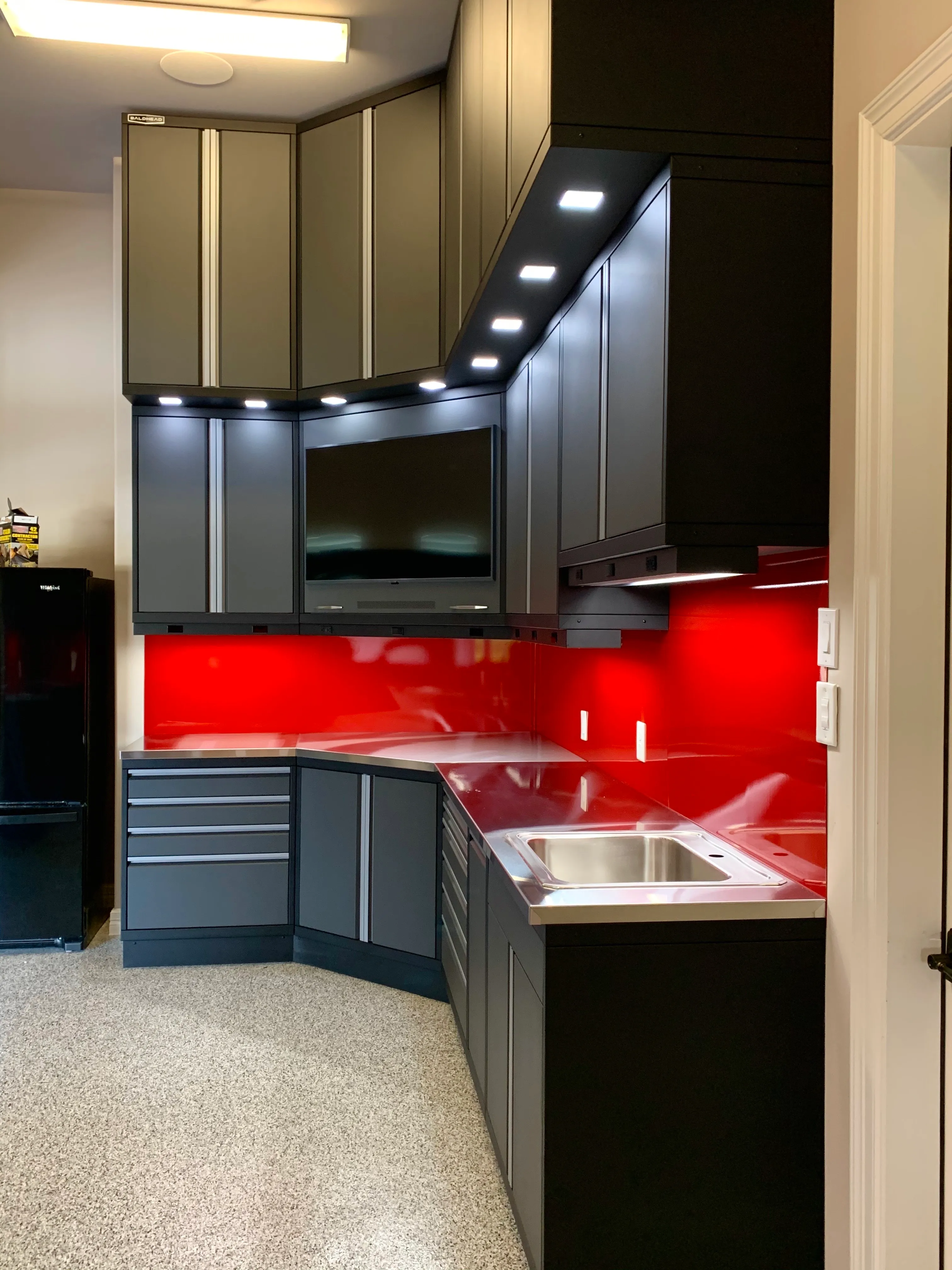 Compact garage workstation by Baldhead Cabinets featuring matte black and gray powder-coated cabinetry, integrated under-cabinet lighting, vivid red high-gloss backsplash, stainless steel countertops and sink, and a wall-mounted TV in a modern epoxy-floored space.
