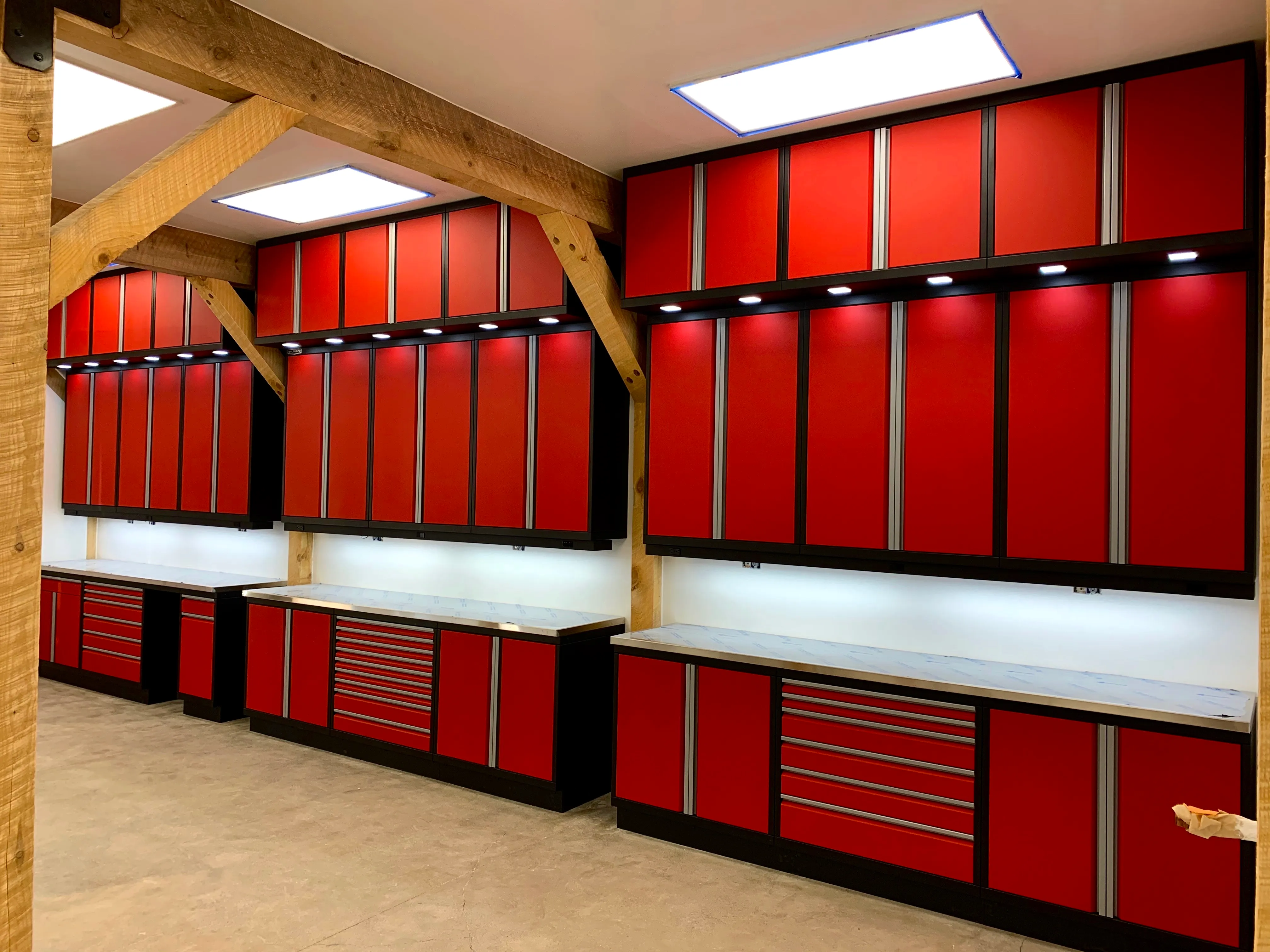 Striking red custom garage cabinets by Baldhead Cabinets featuring full-height upper storage, integrated LED lighting, multiple drawer configurations, and natural wood support beams in a rustic-contemporary workshop environment.