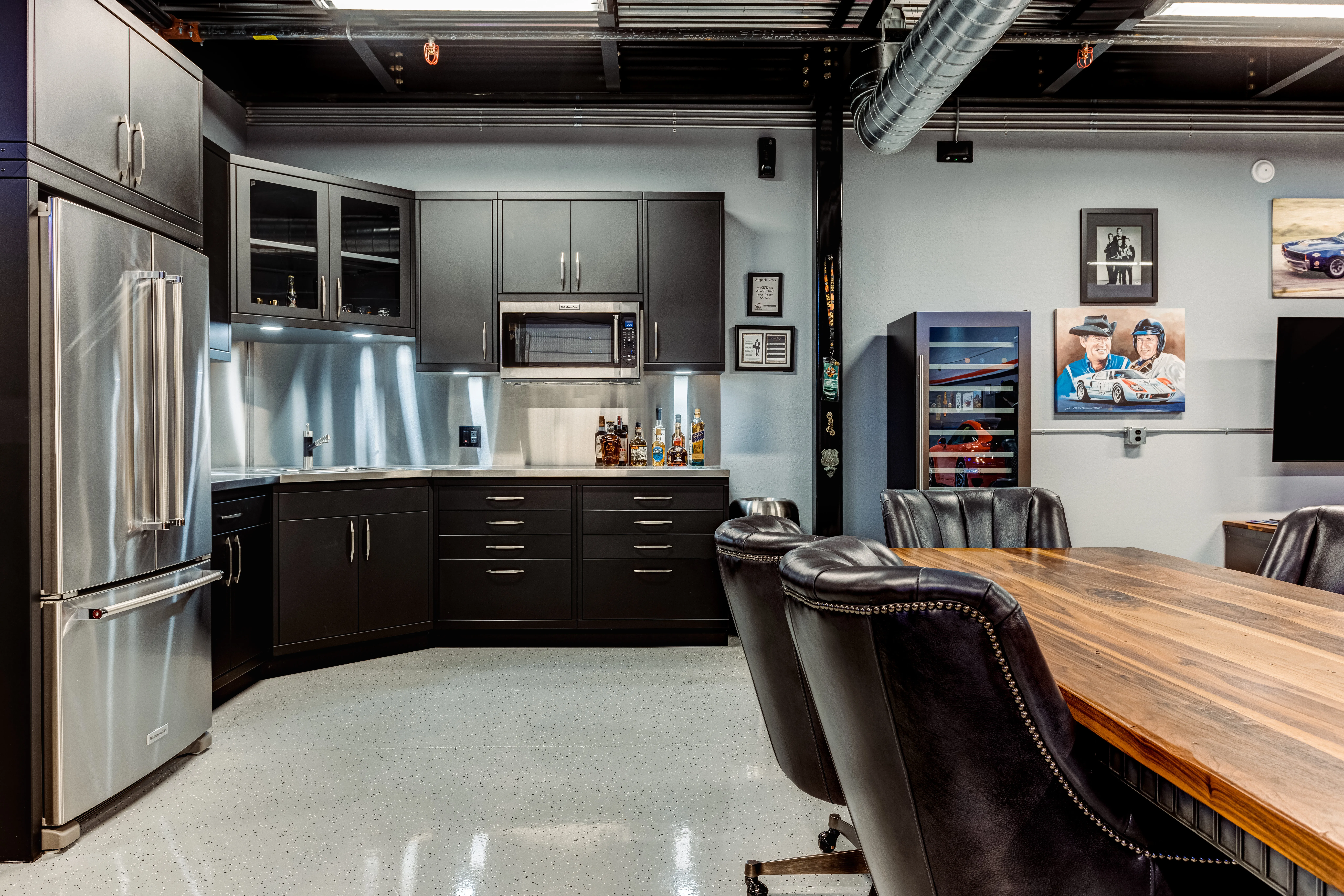 Custom garage lounge and kitchenette design by Baldhead Cabinets, showcasing matte black cabinetry, stainless steel countertops, built-in microwave, under-cabinet lighting, and a stylish meeting table—perfect for blending workspace utility with premium comfort and automotive-themed aesthetics.