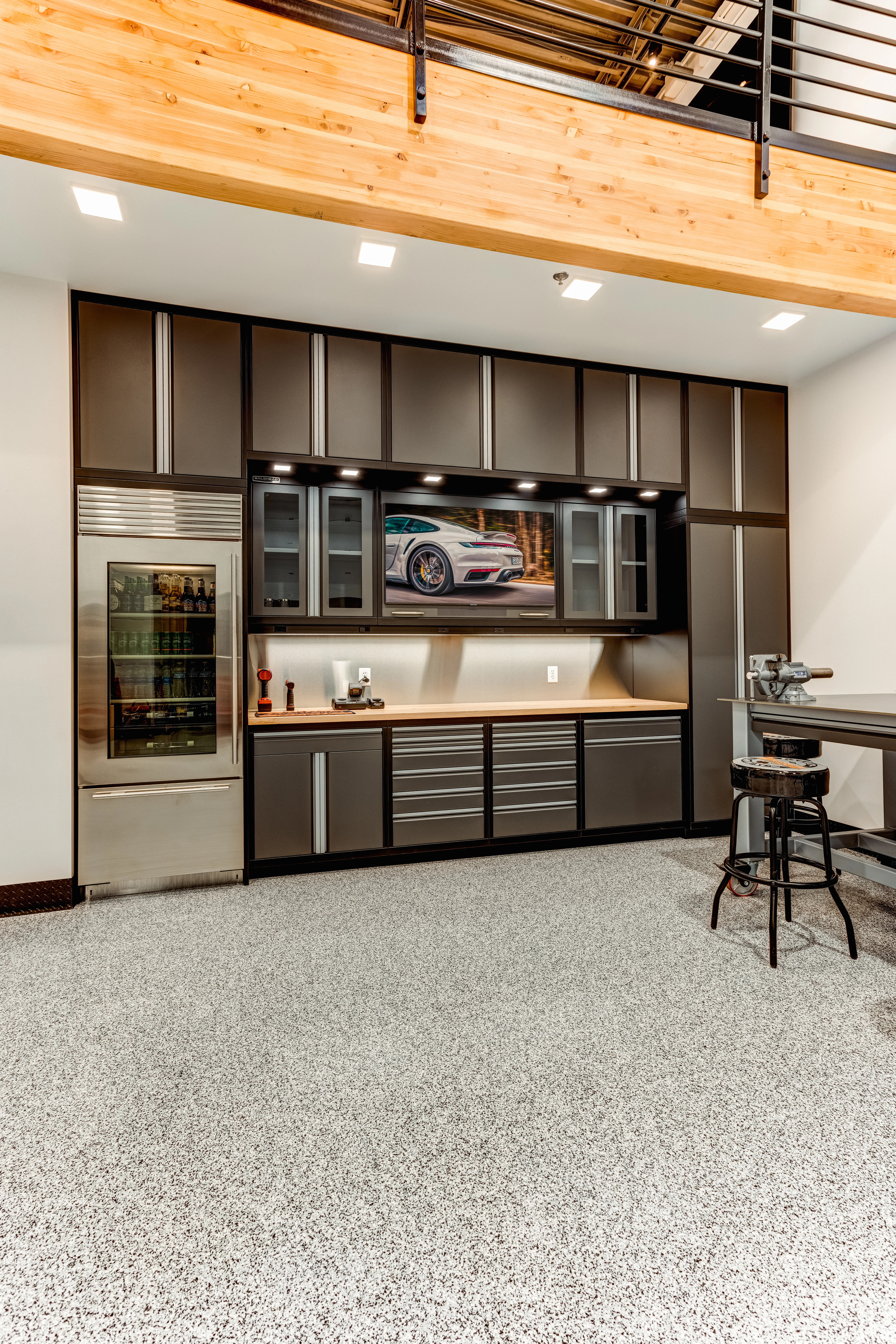Luxury garage cabinetry system by Baldhead Cabinets featuring integrated refrigerator, LED under-cabinet lighting, wood butcher block countertop, upper glass-display cabinets, and a built-in wall-mounted TV — ideal for premium garage storage and workspace solutions.