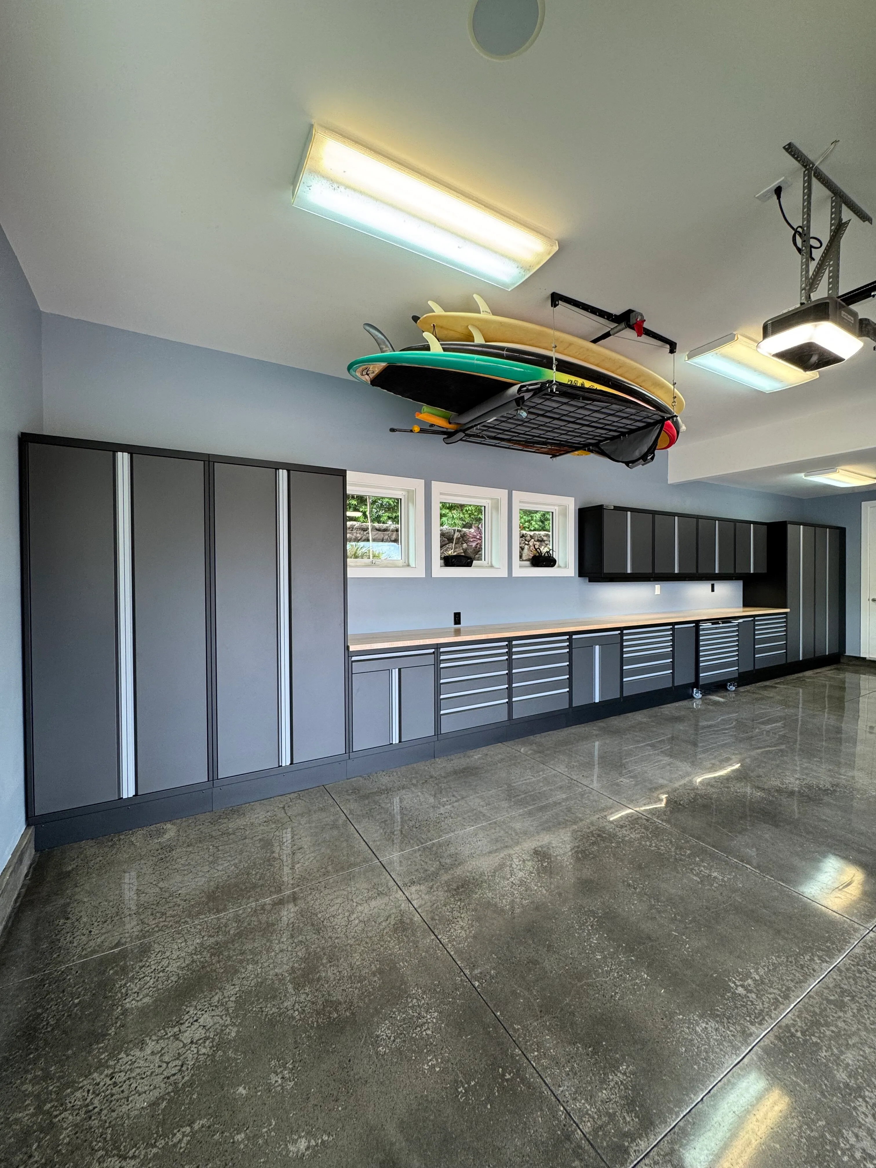 Custom gray garage cabinets by Baldhead Cabinets with butcher block countertops, overhead storage, and integrated drawers, paired with polished concrete floors and ceiling-mounted surfboard rack for a sleek and functional coastal garage setup.