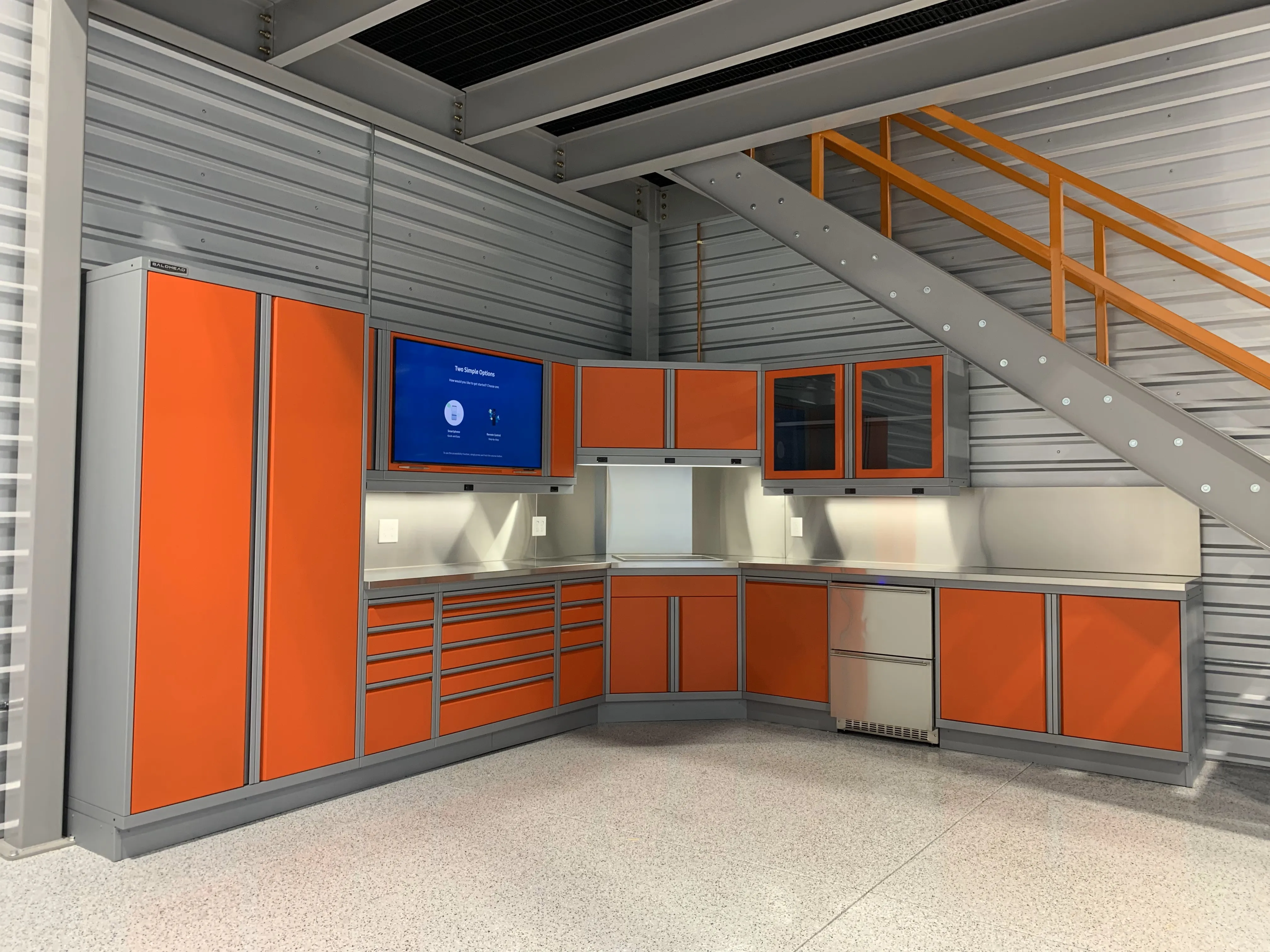 Custom orange garage cabinet system by Baldhead Cabinets, featuring sleek powder-coated steel doors, integrated LED lighting, stainless steel countertops, and wall-mounted storage under a modern staircase in an industrial workshop setting.