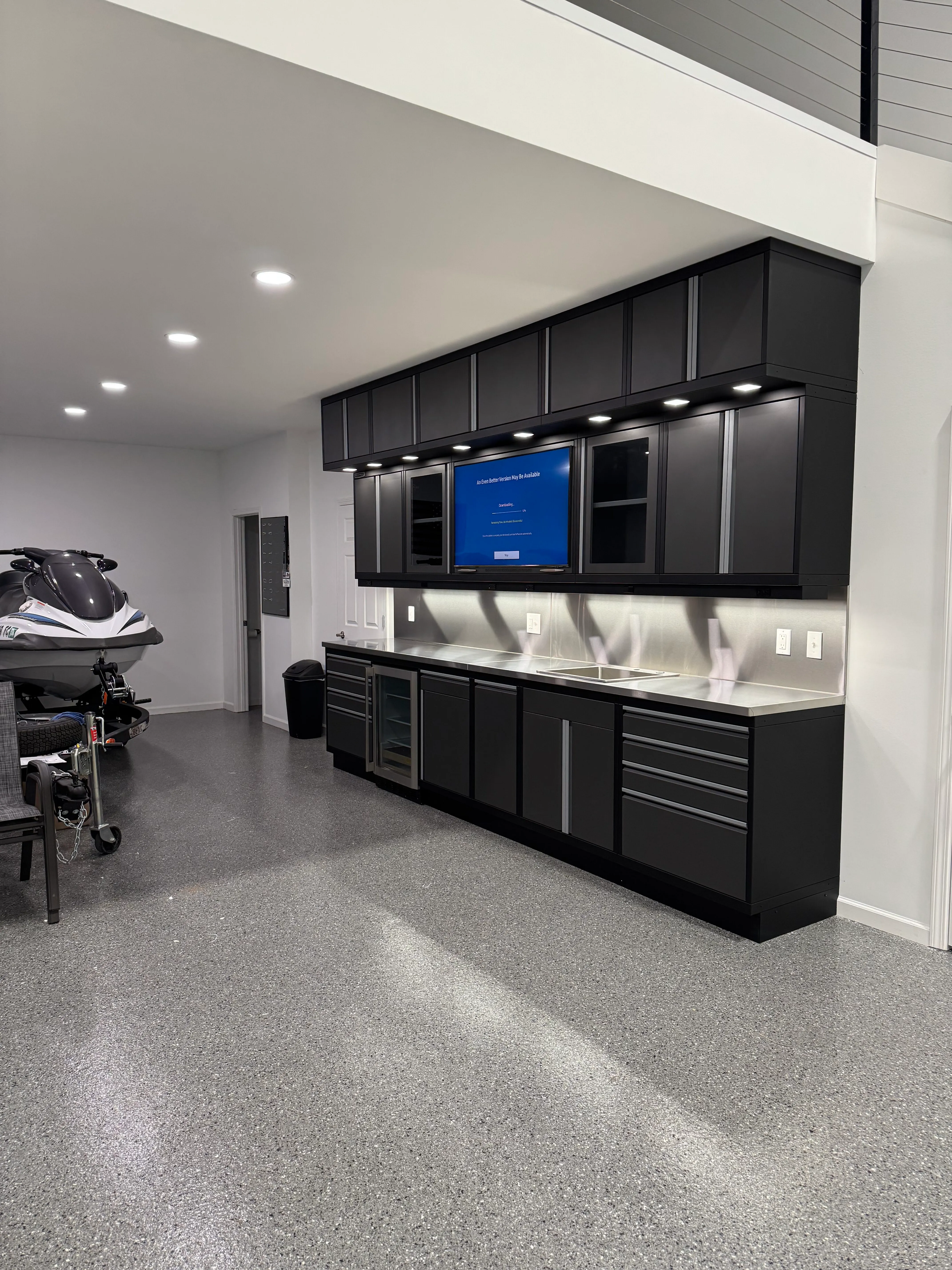 Sleek matte black garage cabinets by Baldhead Cabinets featuring upper and lower storage, stainless steel backsplash, integrated sink, under-cabinet lighting, beverage fridge, wall-mounted TV, and polished epoxy flooring in a luxury marine garage.