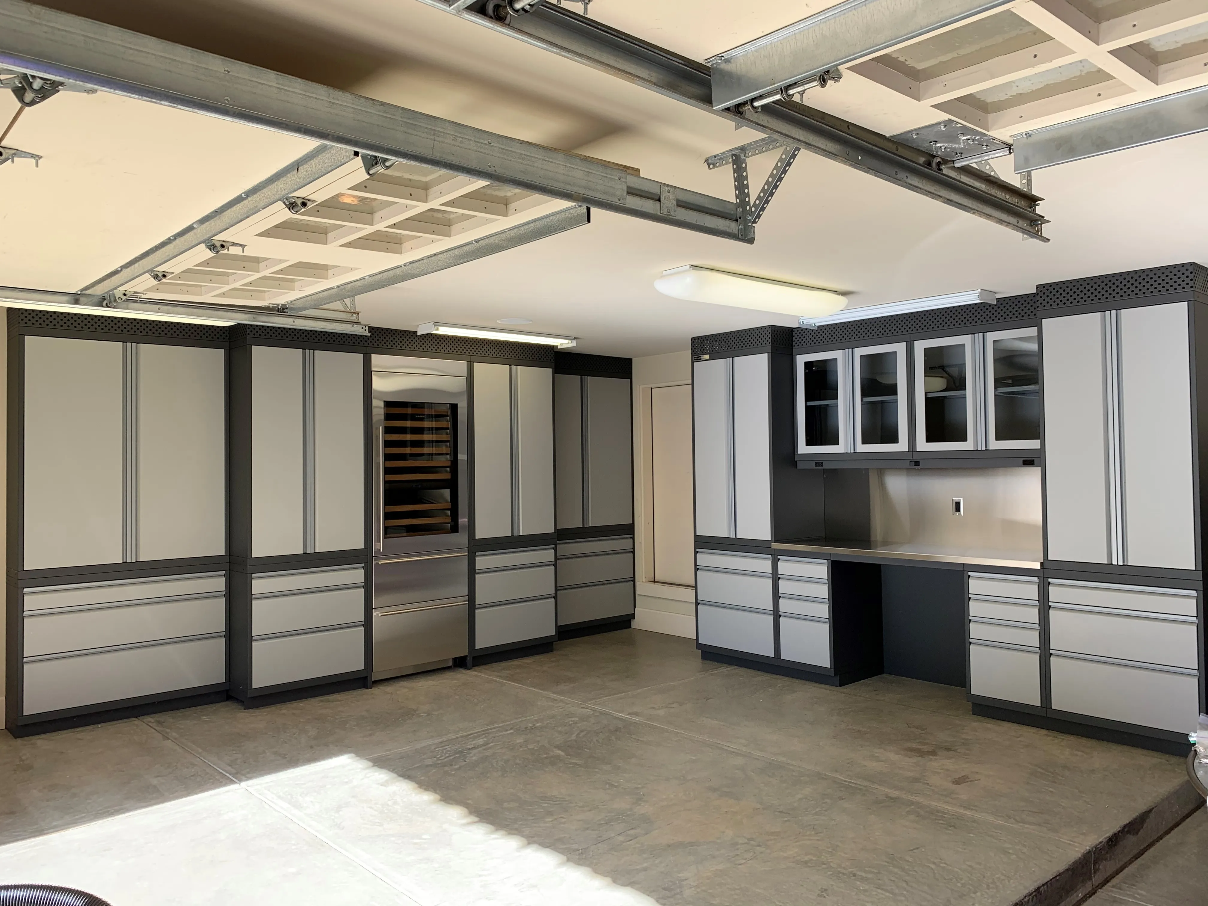 Custom garage storage by Baldhead Cabinets featuring matte gray and black powder-coated cabinetry, glass-front upper cabinets, stainless steel countertops, integrated appliance space, and polished concrete flooring in a luxury home garage setup.