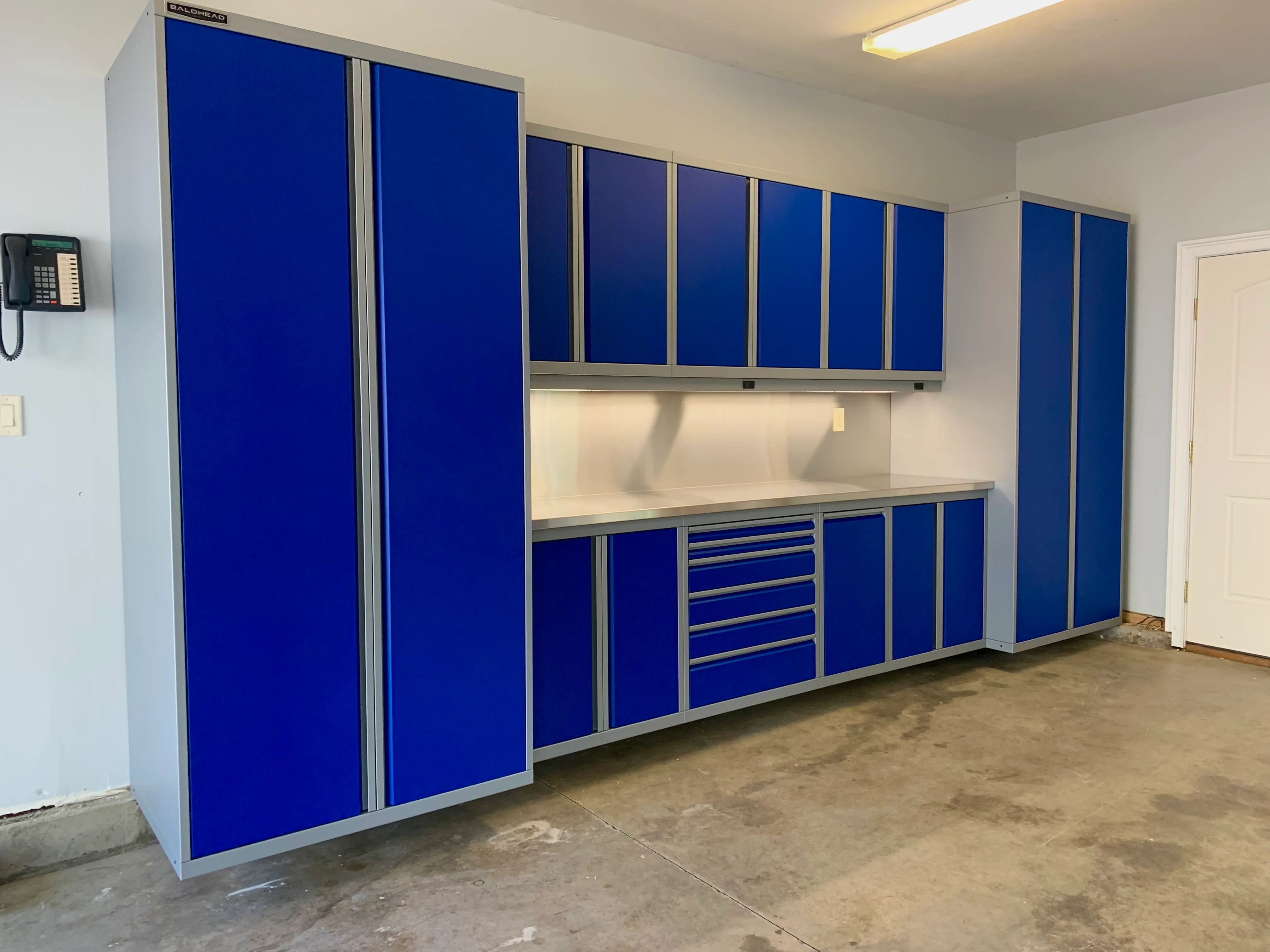 Vibrant blue custom garage cabinets by Baldhead Cabinets featuring tall storage units, upper and lower cabinetry, stainless steel countertop, integrated LED lighting, and sleek aluminum trim in a clean residential garage workspace.