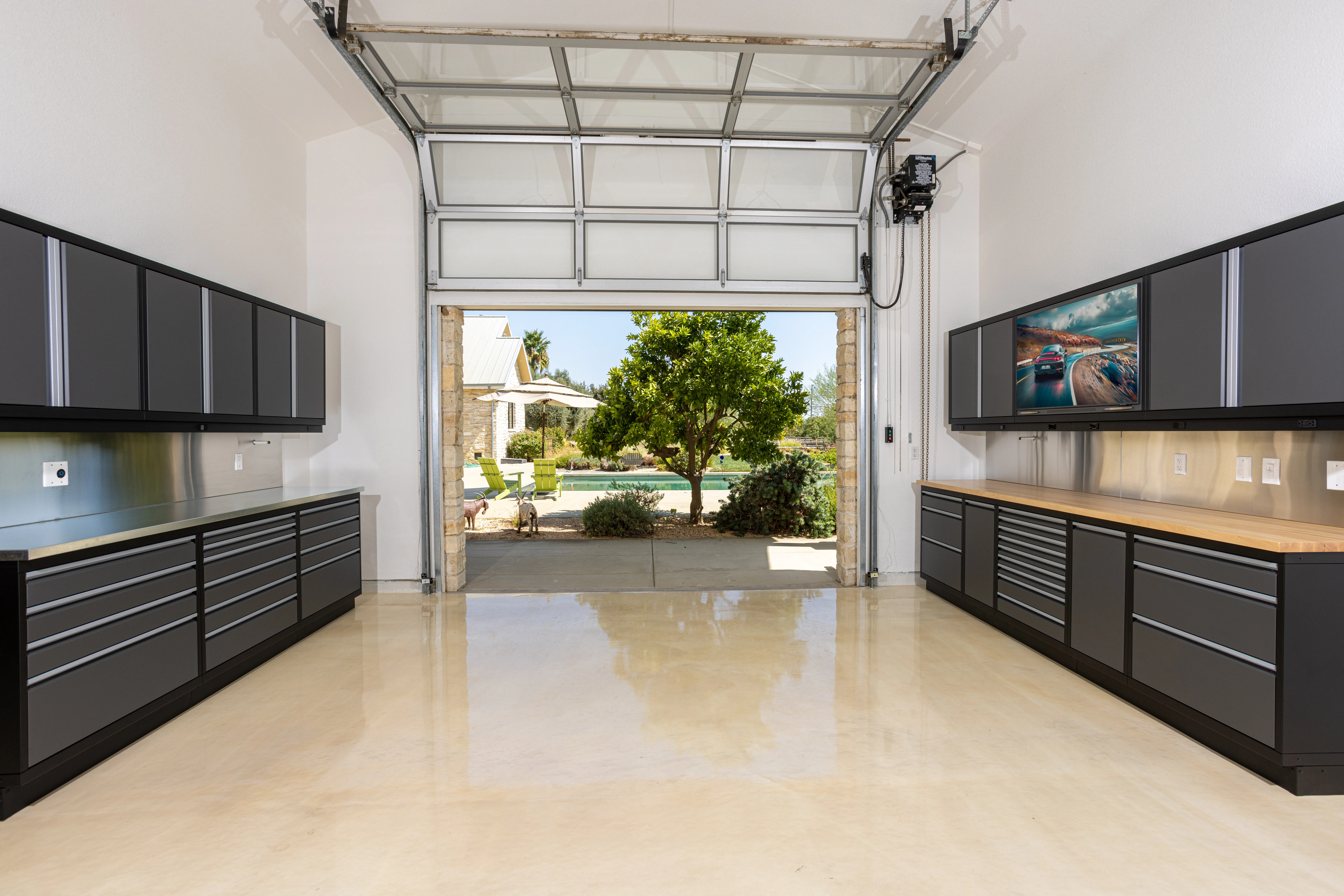 Custom garage workspace with dual wall-mounted cabinet systems by Baldhead Cabinets, featuring stainless steel countertops, matte black finish, and polished concrete flooring—designed for high-end garage organization with a clean, open view to a landscaped backyard.