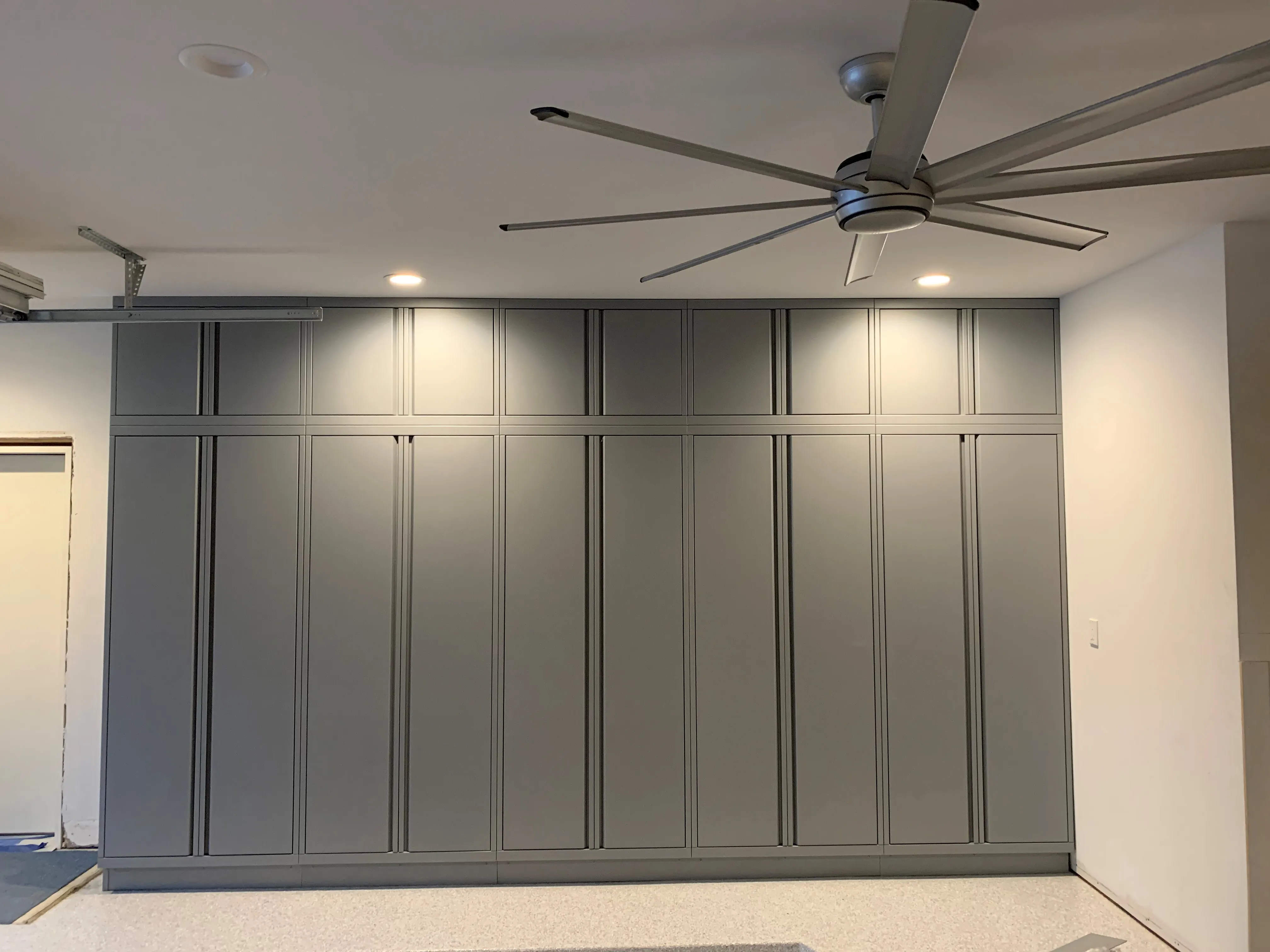 Sleek wall-to-wall garage storage by Baldhead Cabinets featuring tall matte gray powder-coated cabinets with minimalist handles, recessed lighting, and a large industrial ceiling fan, providing clean, modern organization for a high-end garage.