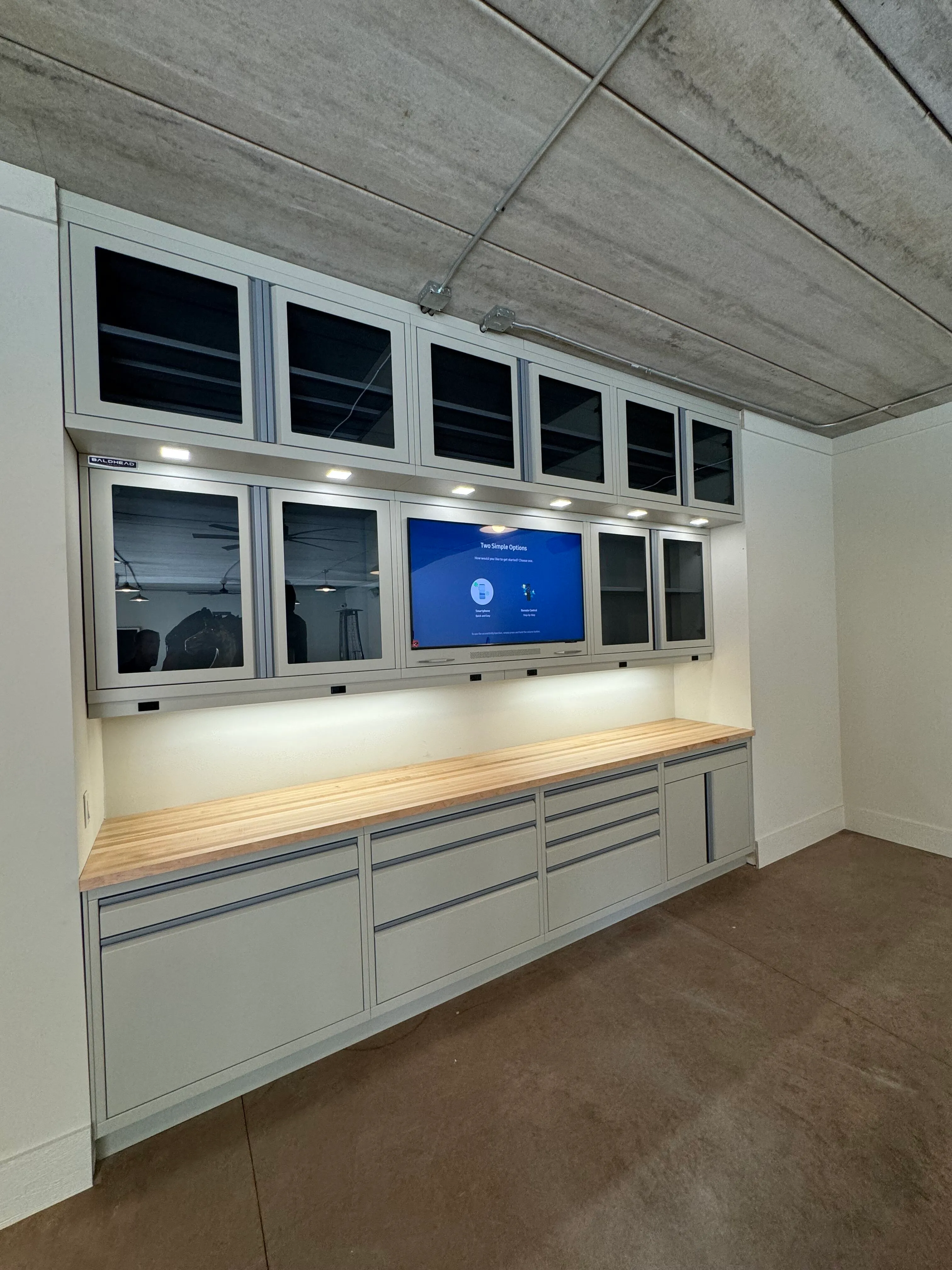 Custom wall-mounted garage cabinetry system by Baldhead Cabinets featuring glass-front upper cabinets, built-in lighting, wood butcher block countertop, and integrated television — ideal for modern, high-functioning garage workspaces.