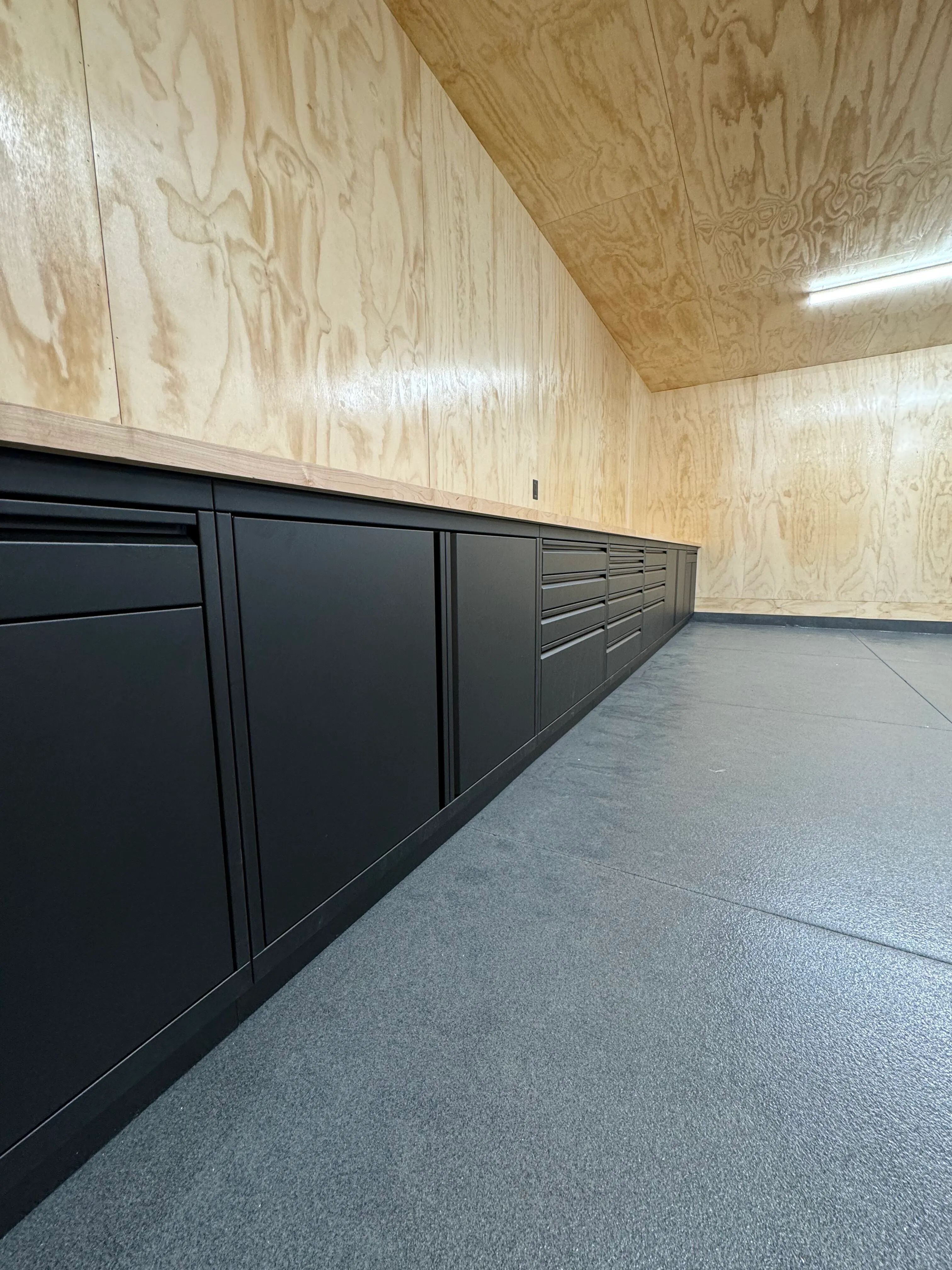 Matte black custom lower garage cabinets by Baldhead Cabinets, installed against natural plywood walls with wood countertop and textured epoxy flooring, creating a minimalist, modern workshop with durable storage solutions.