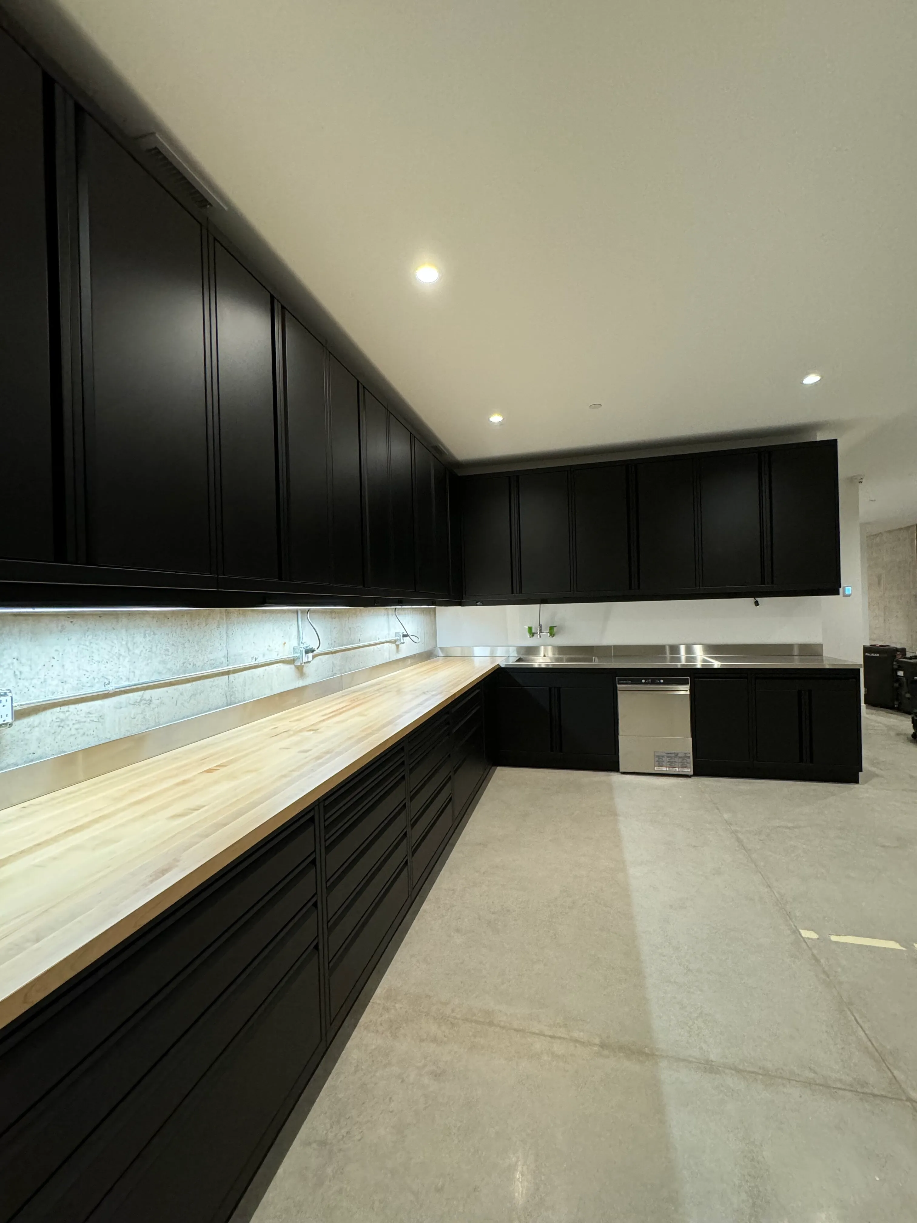 Modern matte black custom garage cabinets by Baldhead Cabinets, featuring full-length butcher block and stainless steel countertops, upper and lower storage solutions, integrated dishwasher, and clean concrete flooring in a premium workshop space.