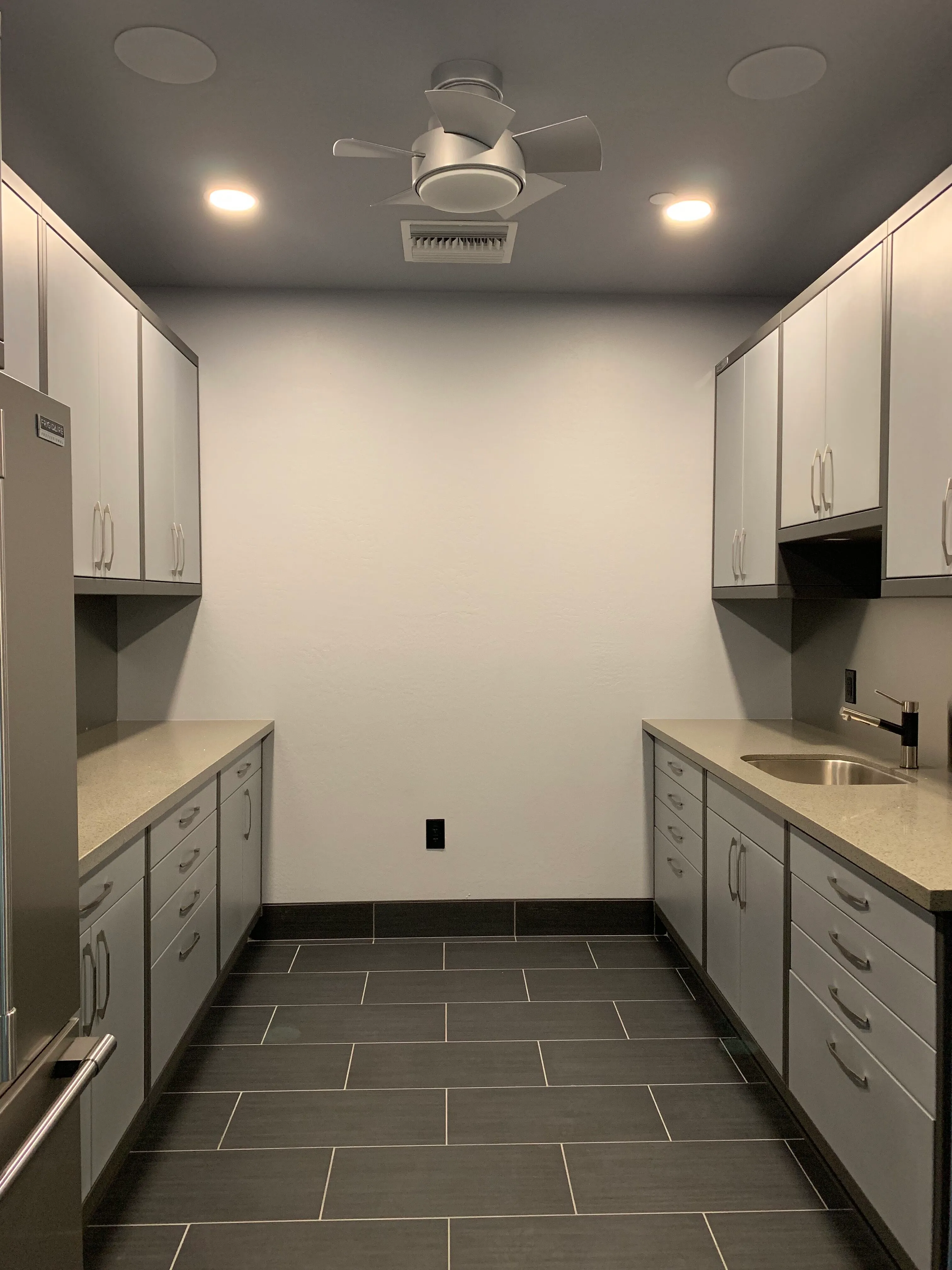 Contemporary utility room with custom cabinetry by Baldhead Cabinets, featuring sleek gray upper and lower storage, quartz countertops, integrated sink, modern ceiling fan, and dark tile flooring for a clean and functional workspace.