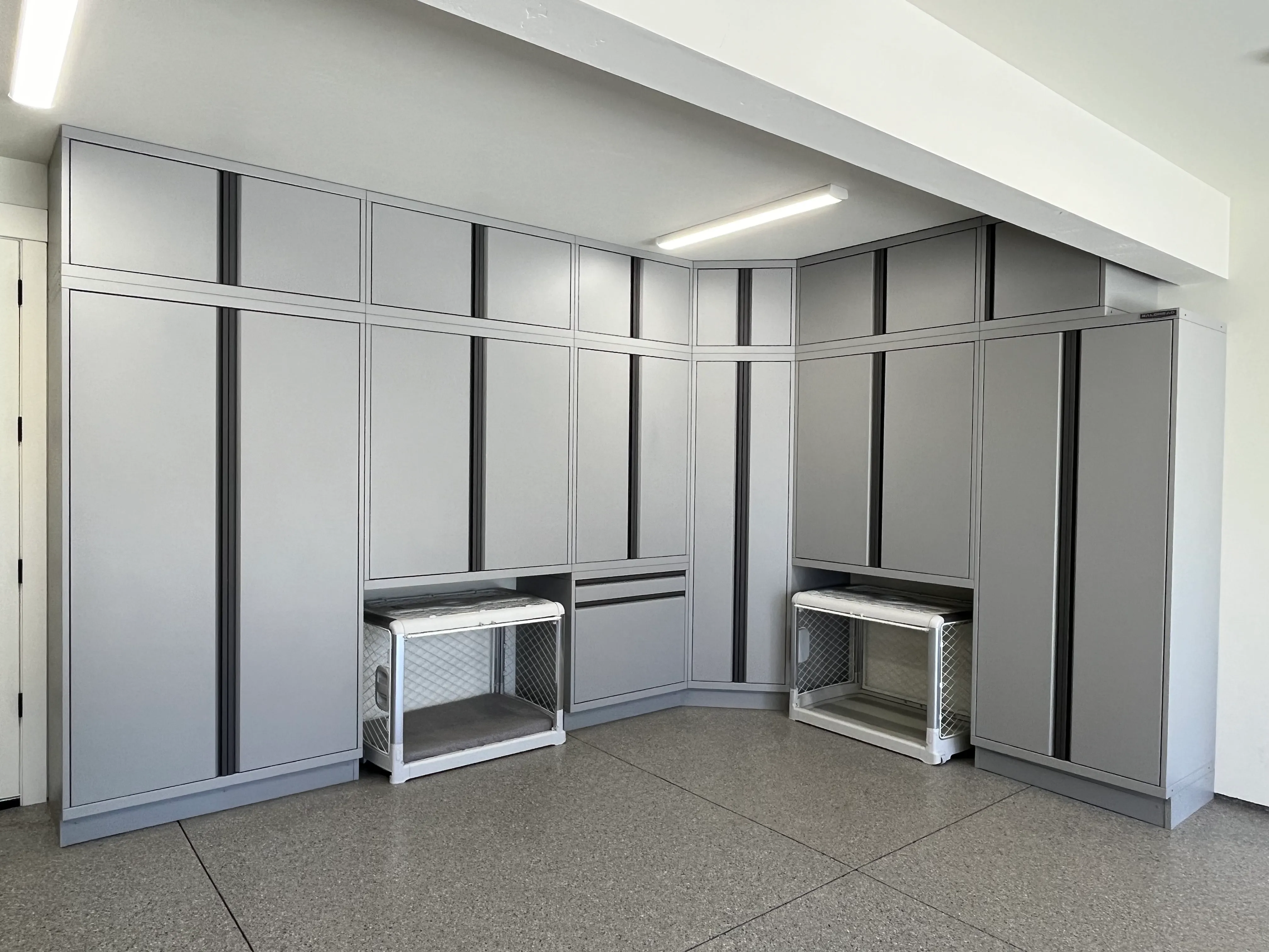 Custom garage corner setup with sleek light gray Baldhead Cabinets, integrated wall-to-wall storage, vertical aluminum trim accents, and built-in cubby spaces designed for pet crates or utility use—ideal for modern garage organization and space optimization.